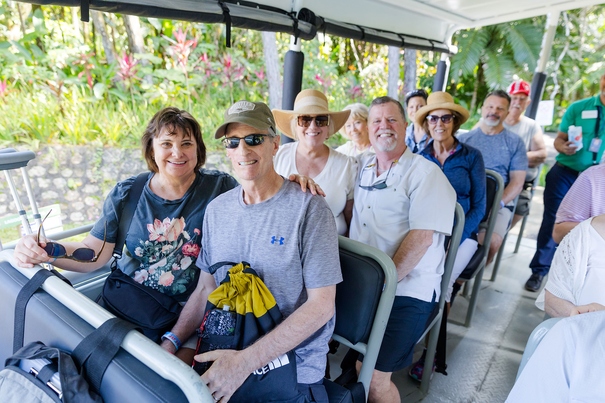 A group of travelers seated in a tour bus, smiling and enjoying the journey, with vibrant tropical plants visible in the background.