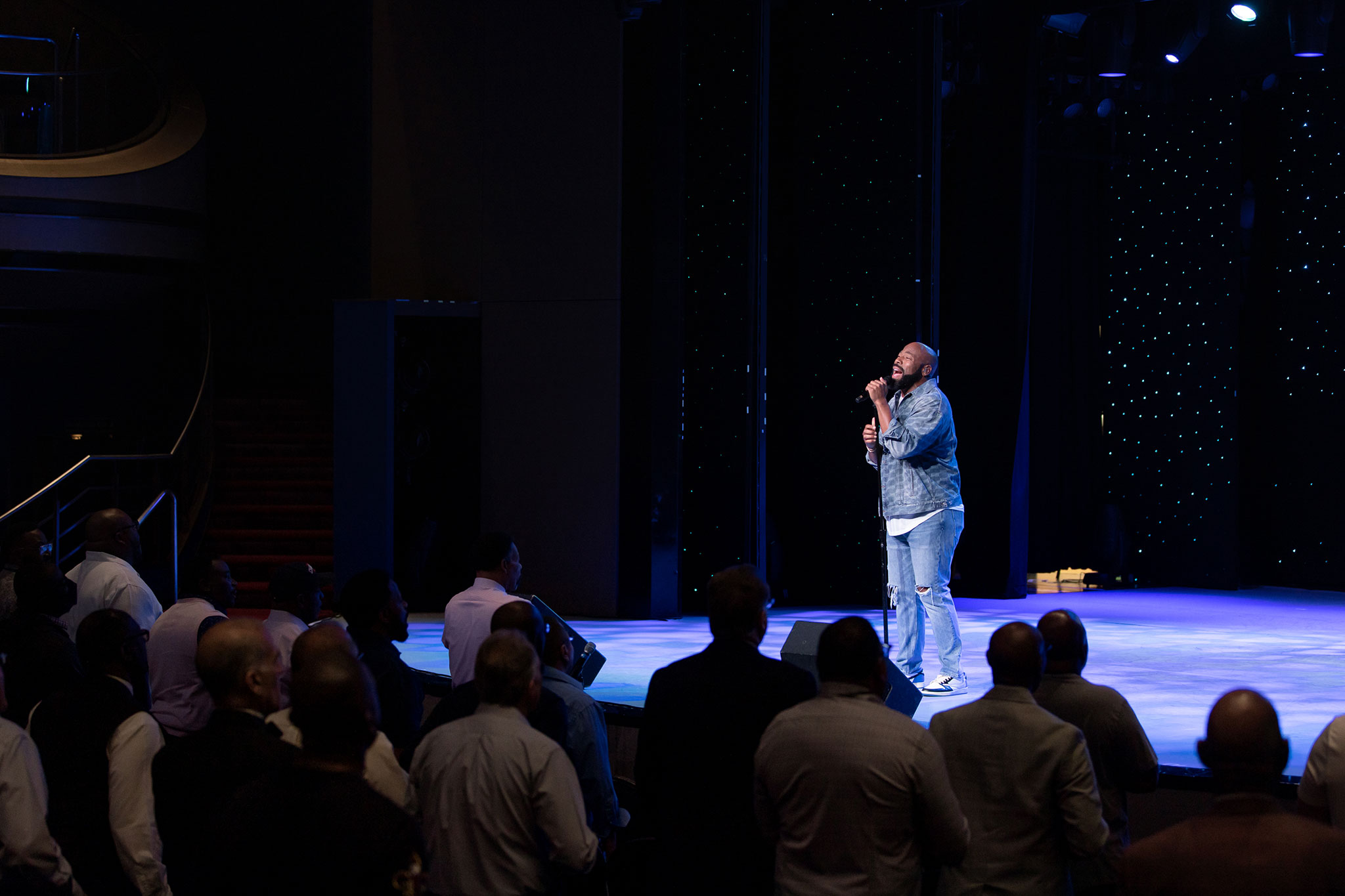 A vocalist performing solo on a stage with a starry backdrop, singing to a crowd of seated attendees.
