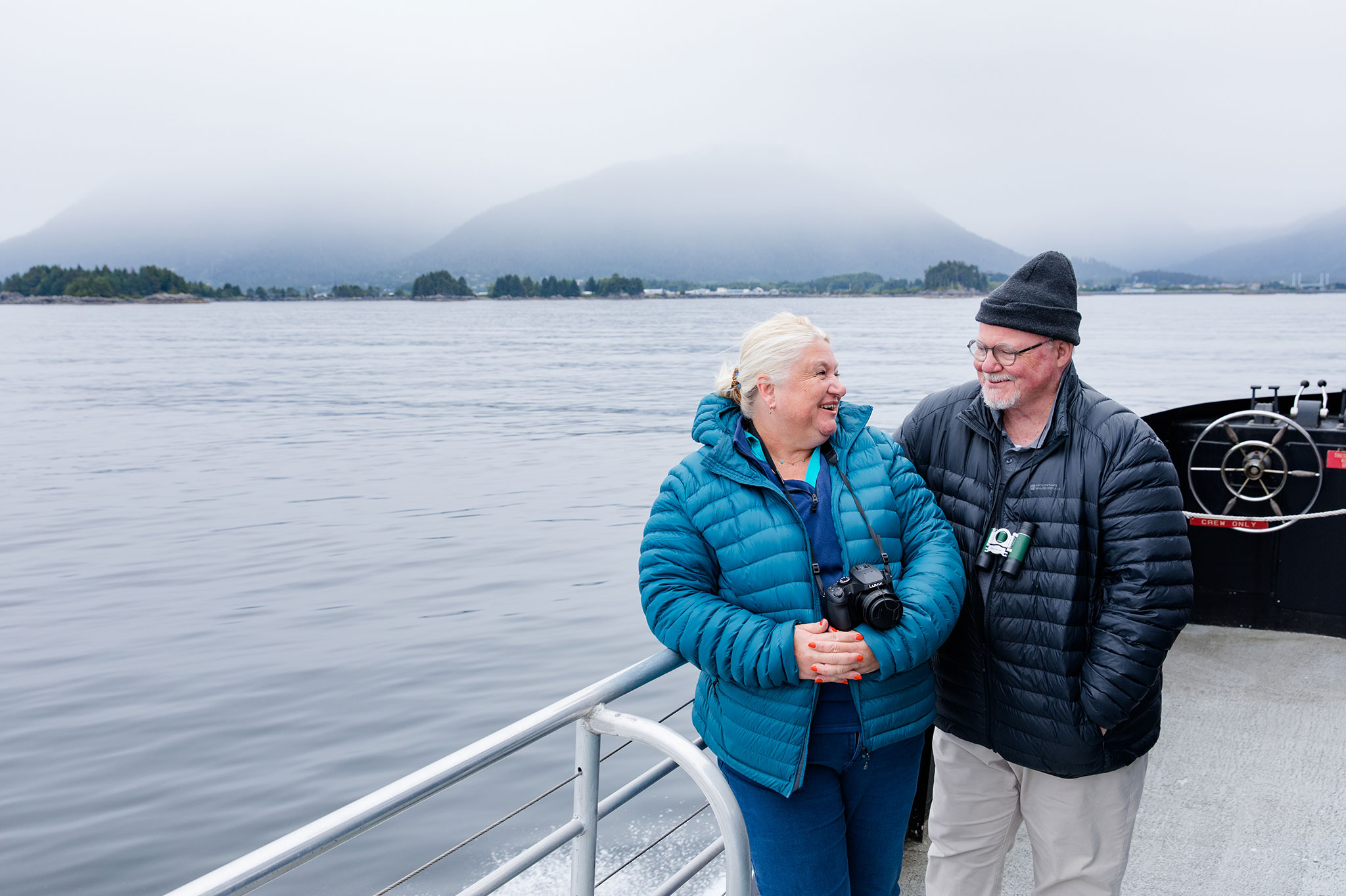 An elderly couple smiling and chatting on a boat with misty mountains in the background.