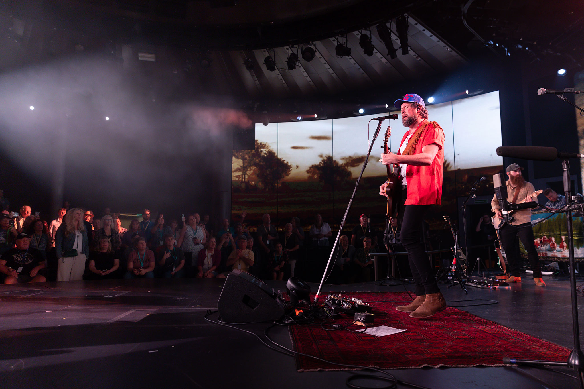 Zach Williams in a red shirt and black pants playing guitar and singing onstage, with an audience in the background.