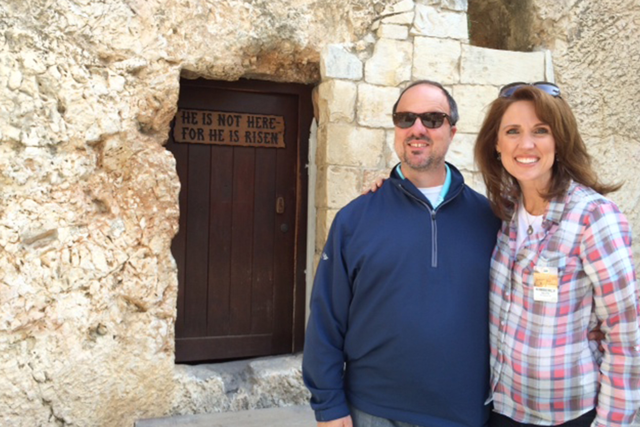 Man and woman smiling in front of the Garden Tomb in Jerusalem, in front of the sign that reads “He is not here—for He is risen.”
