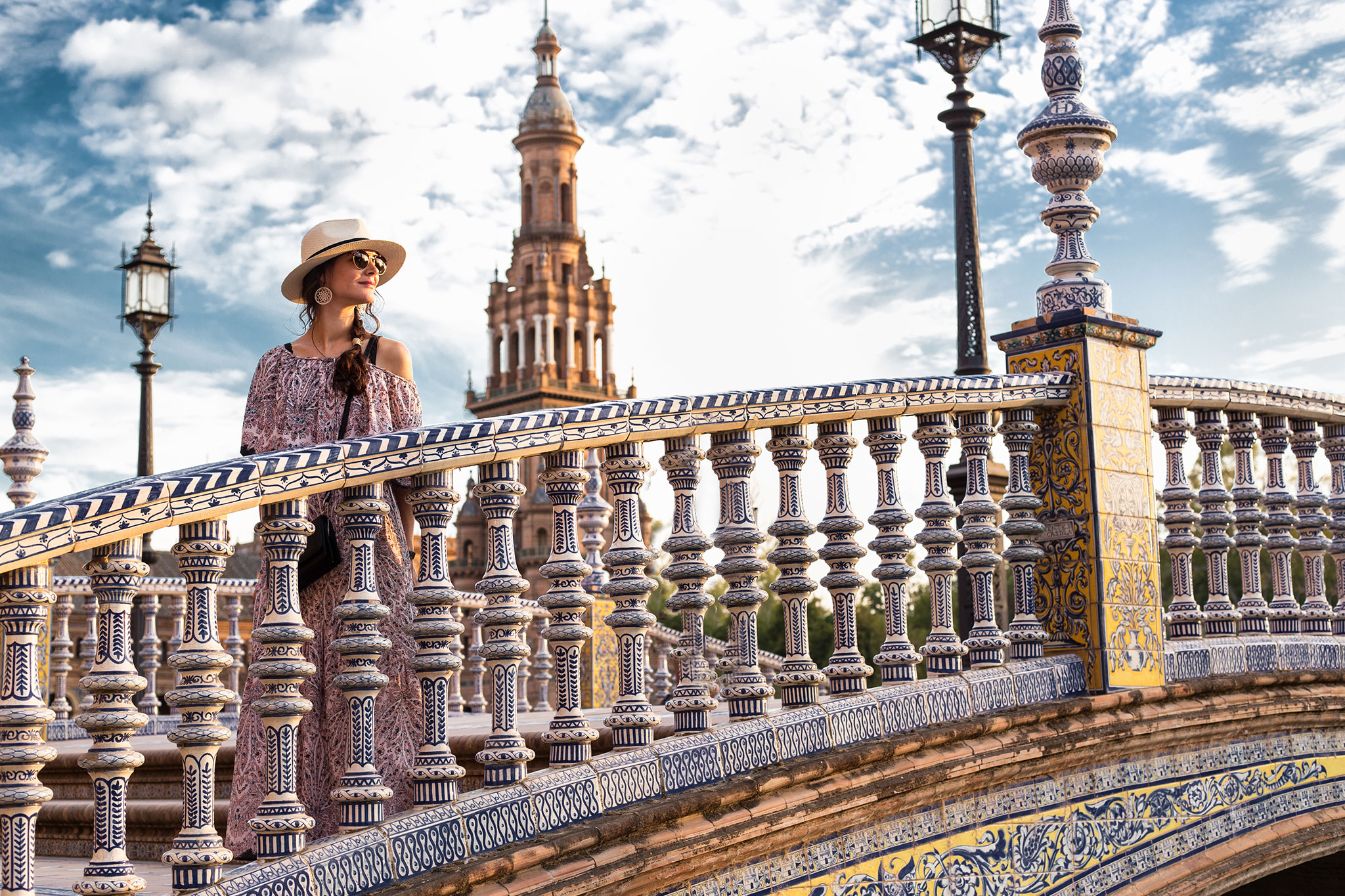 A woman in a hat stands on a colorful tiled bridge in Seville.