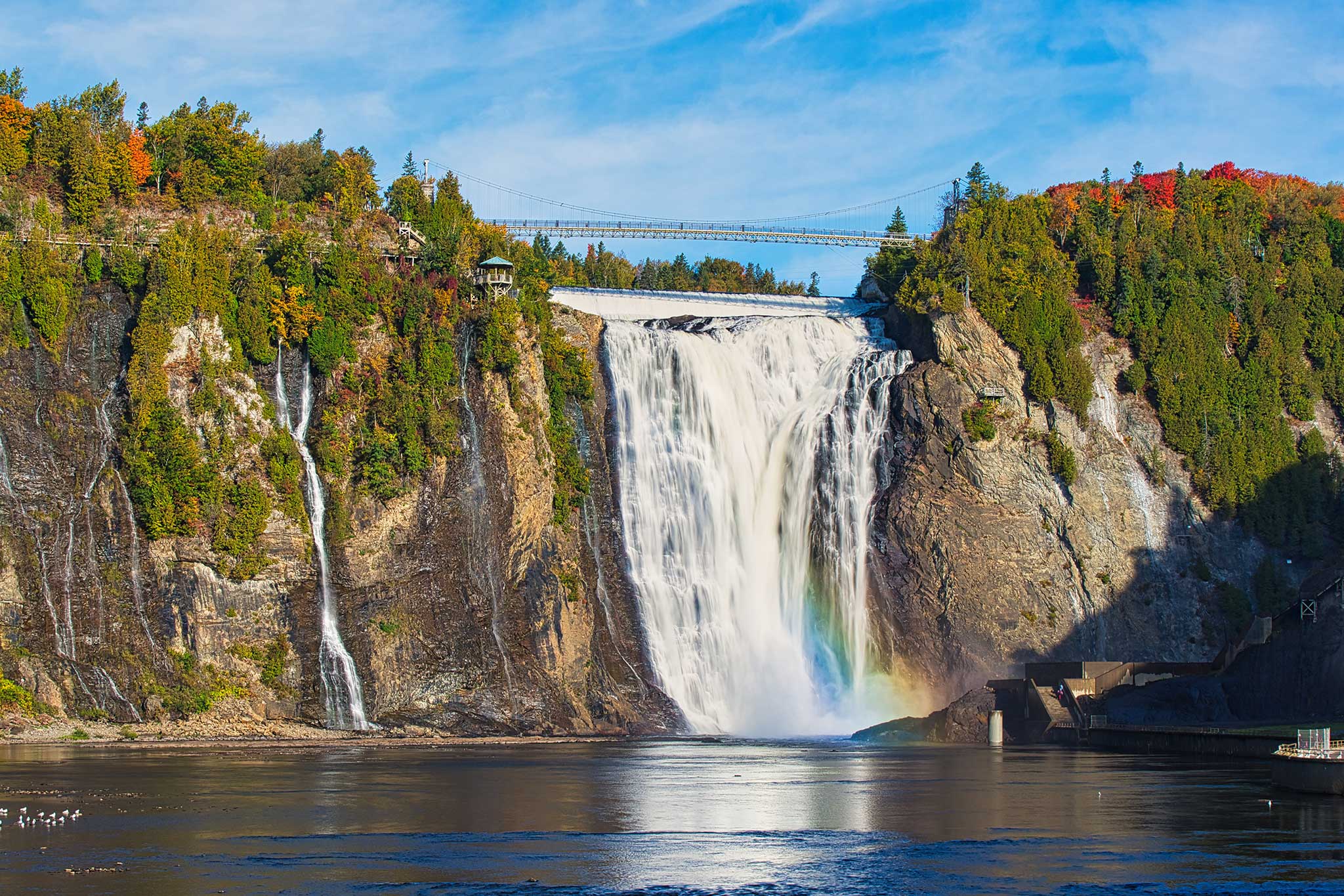 Powerful waterfall cascading down a rocky cliff into a misty river, with a bridge spanning overhead.