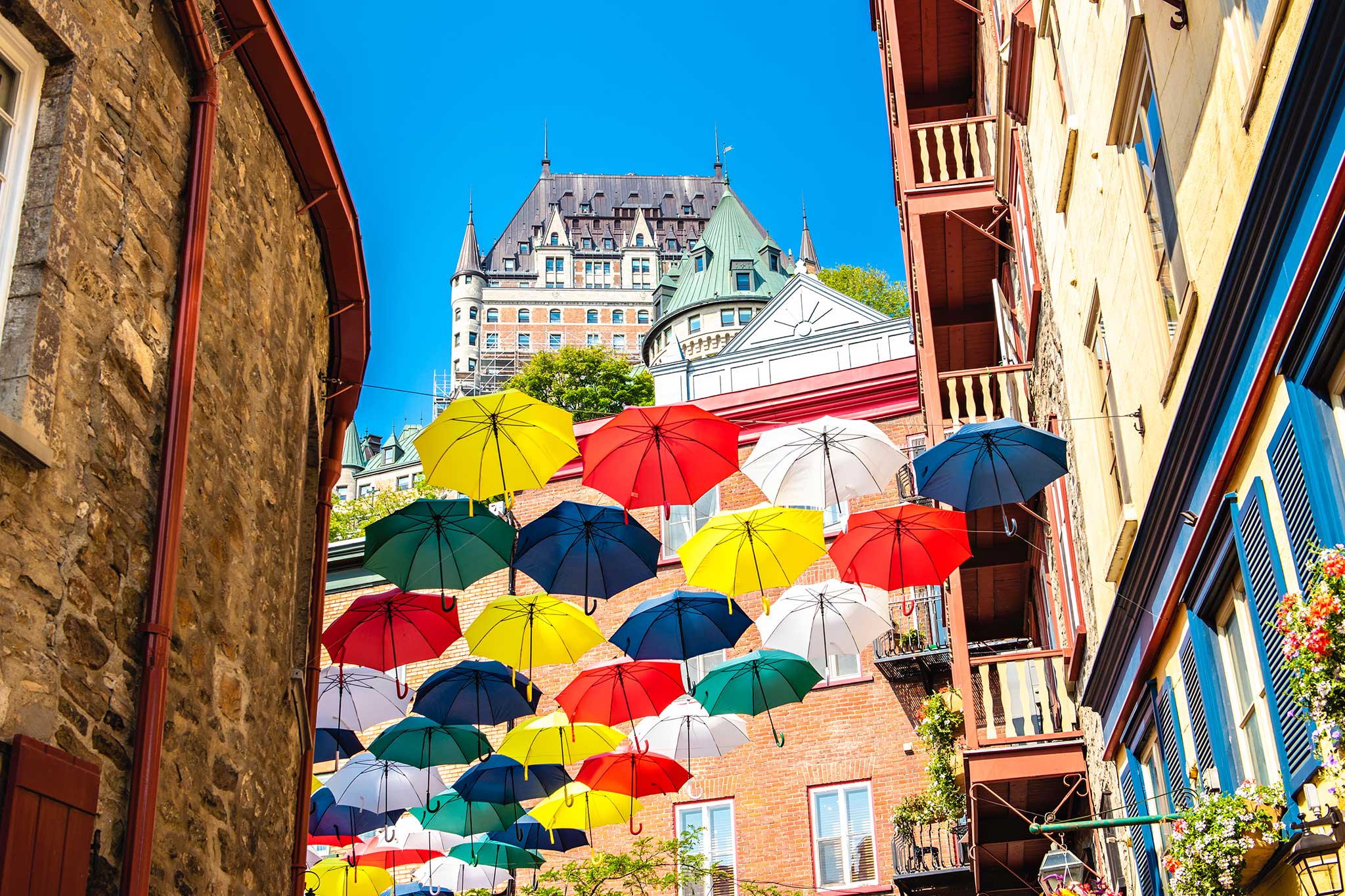 Colorful umbrellas hang above a narrow street in Old Quebec with Chateau Frontenac in the background.