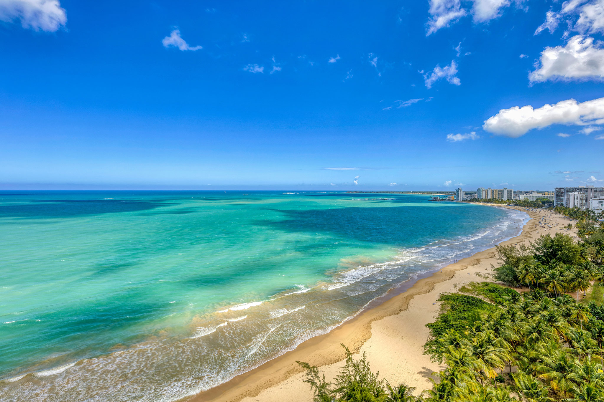 Aerial view of Isla Verde Beach with turquoise waters and clear waves lapping against a sandy shoreline, stretching toward a distant city skyline and lush greenery.
