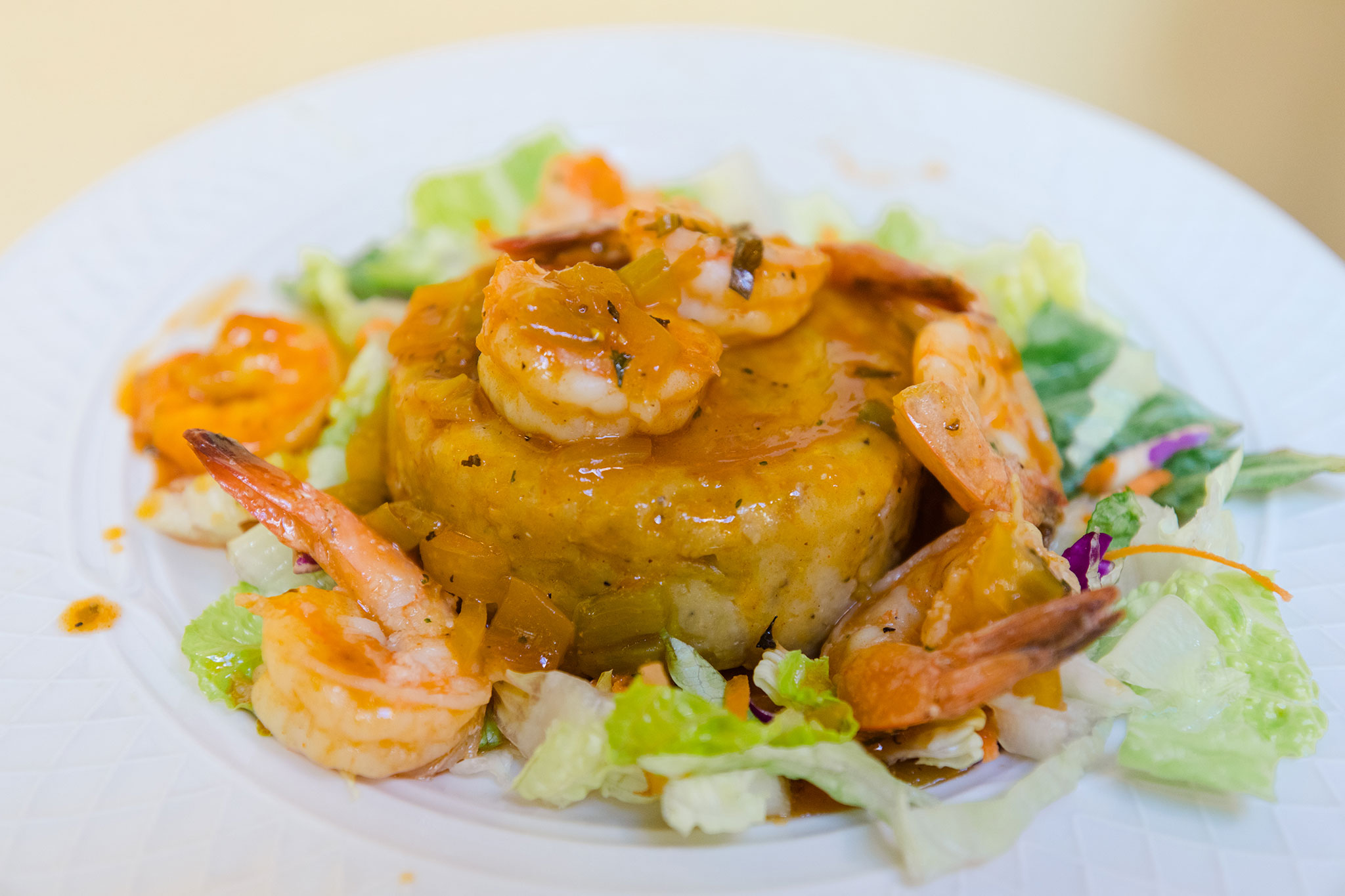 Golden mashed plantains formed into a mound, covered in garlic shrimp and rich orange sauce, with a side of crisp greens.