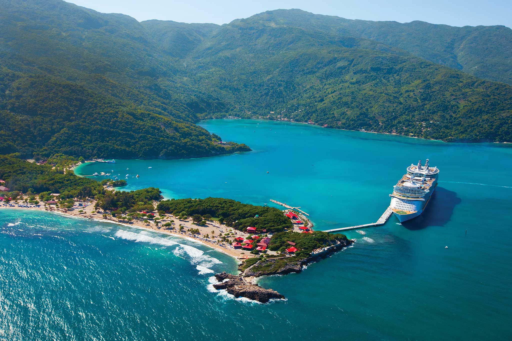 Aerial view of a cruise ship docked at a tropical island with clear turquoise water, golden beaches, a vibrant beachside village, and lush green mountains in the background.