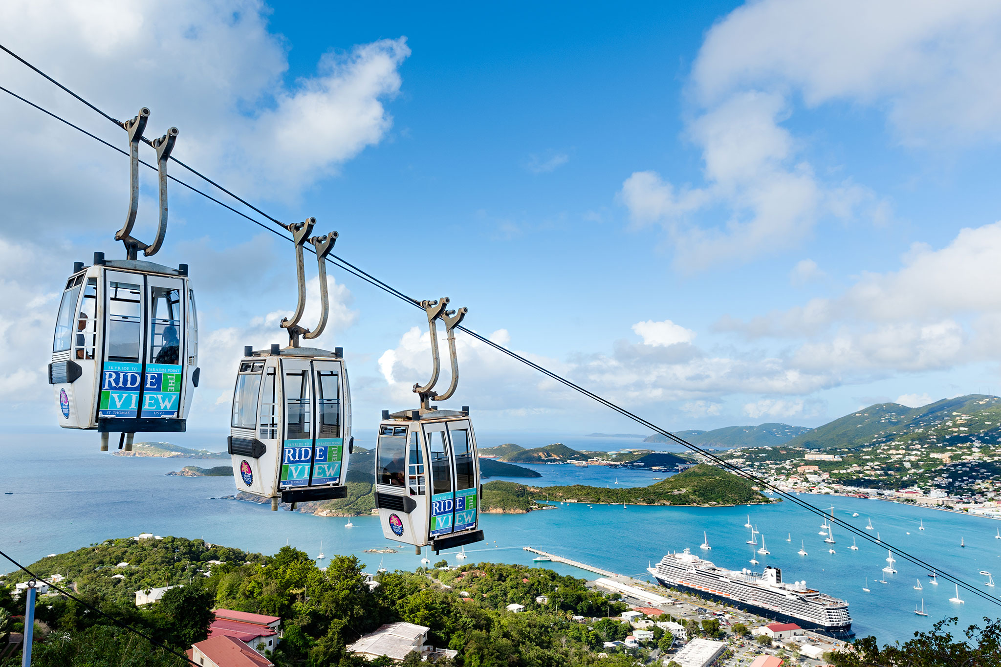 Scenic cable cars glide above a tropical island with views of turquoise bays and a docked cruise ship.