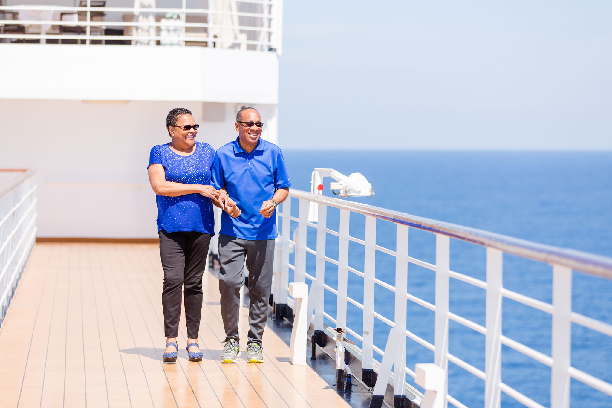 A smiling older couple walks arm in arm along a sunny cruise ship deck with the ocean in the background.