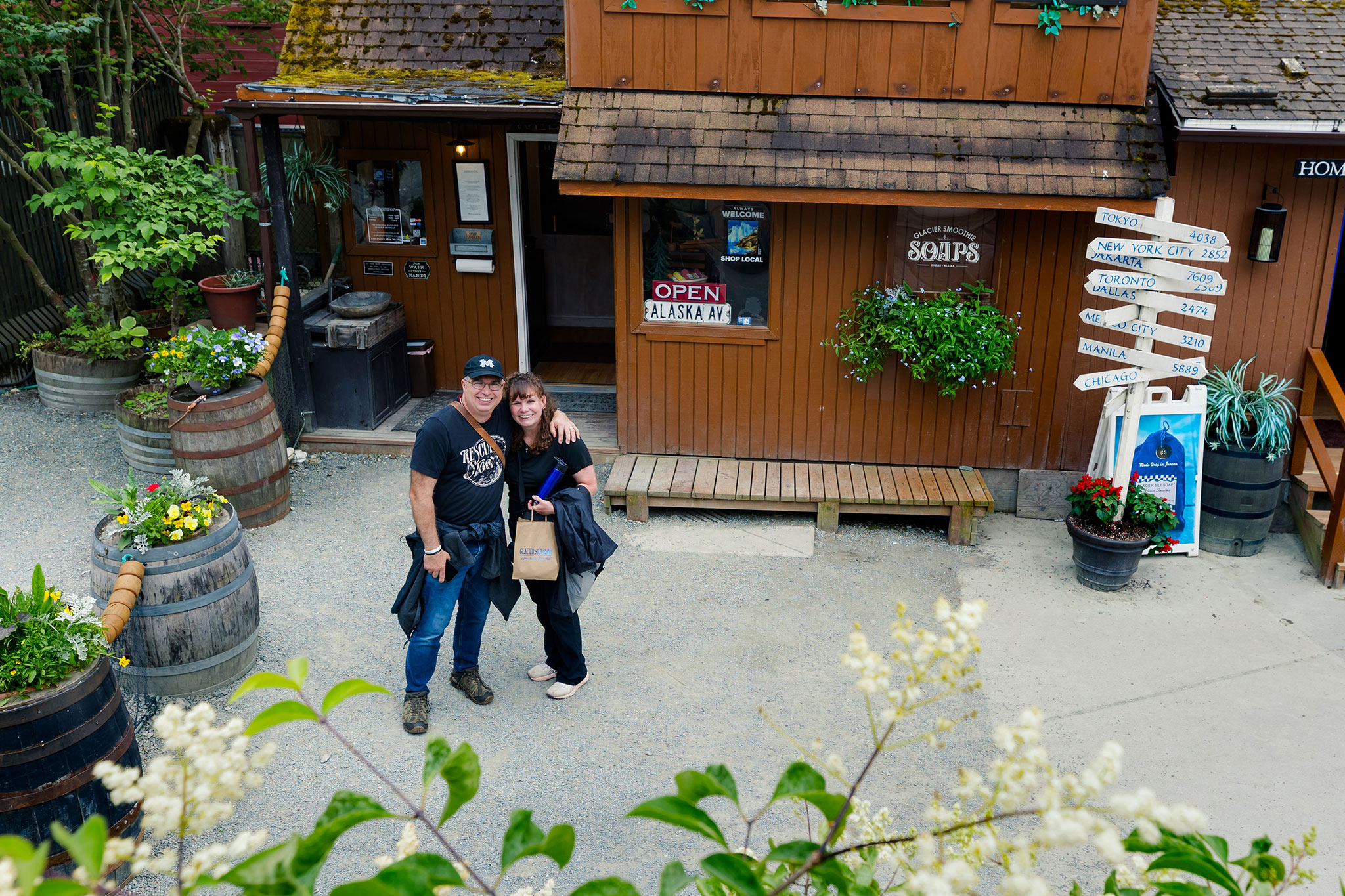 A man and a woman pose together outside a wooden storefront in Alaska, surrounded by plants and signs.