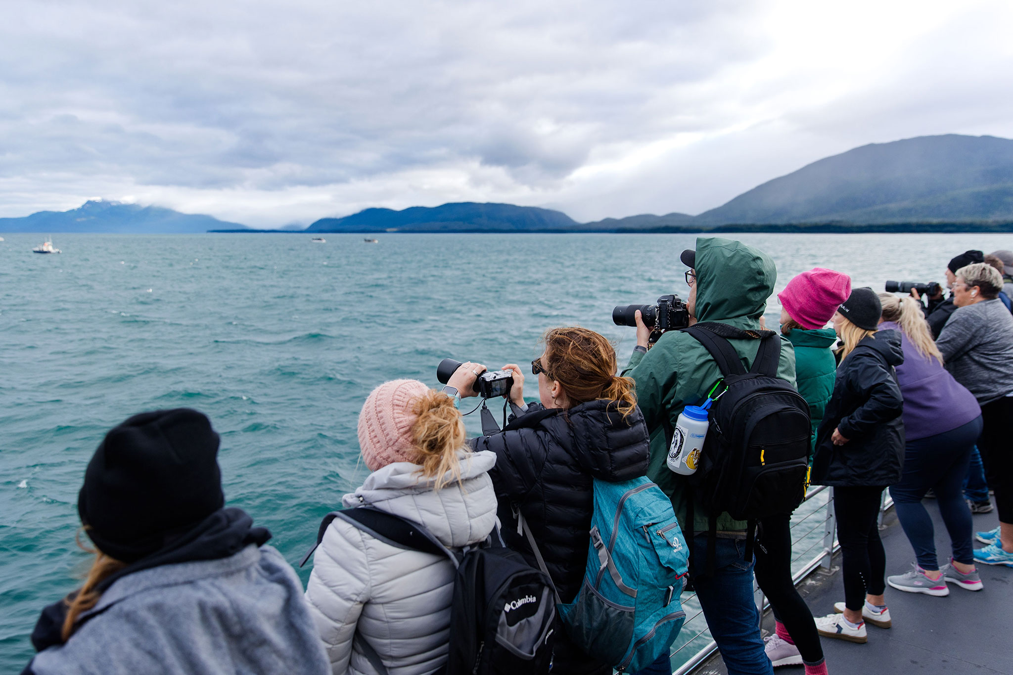 Bundled-up travelers line the railing of a boat, taking photos of the scenic ocean and distant mountains under cloudy skies.