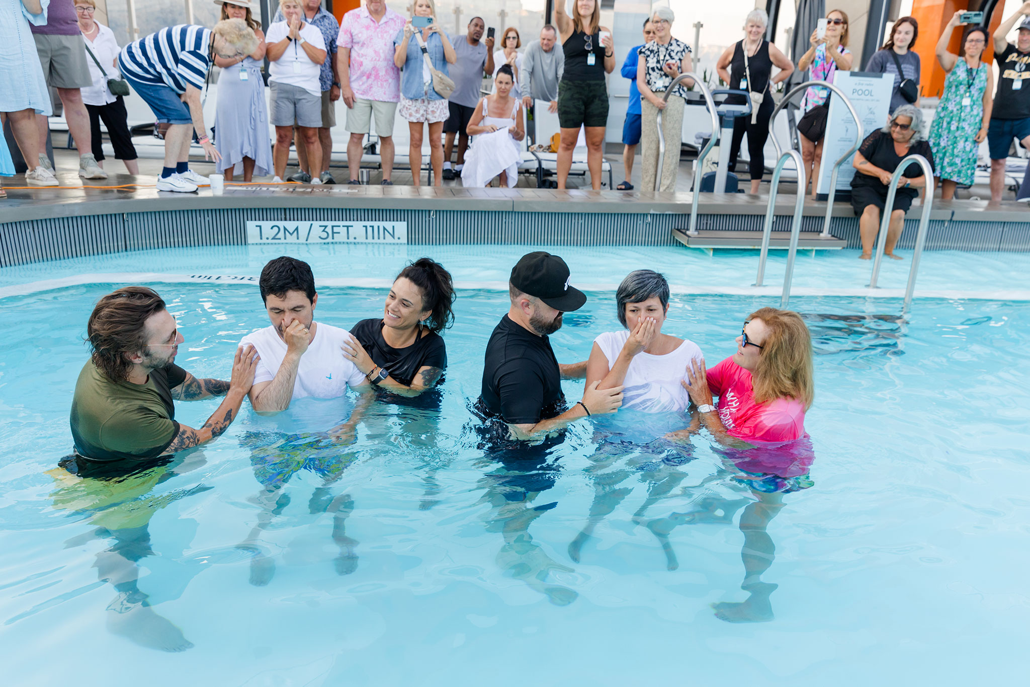 An impromptu baptism ceremony takes place in a cruise ship pool as a group of people watch from the deck