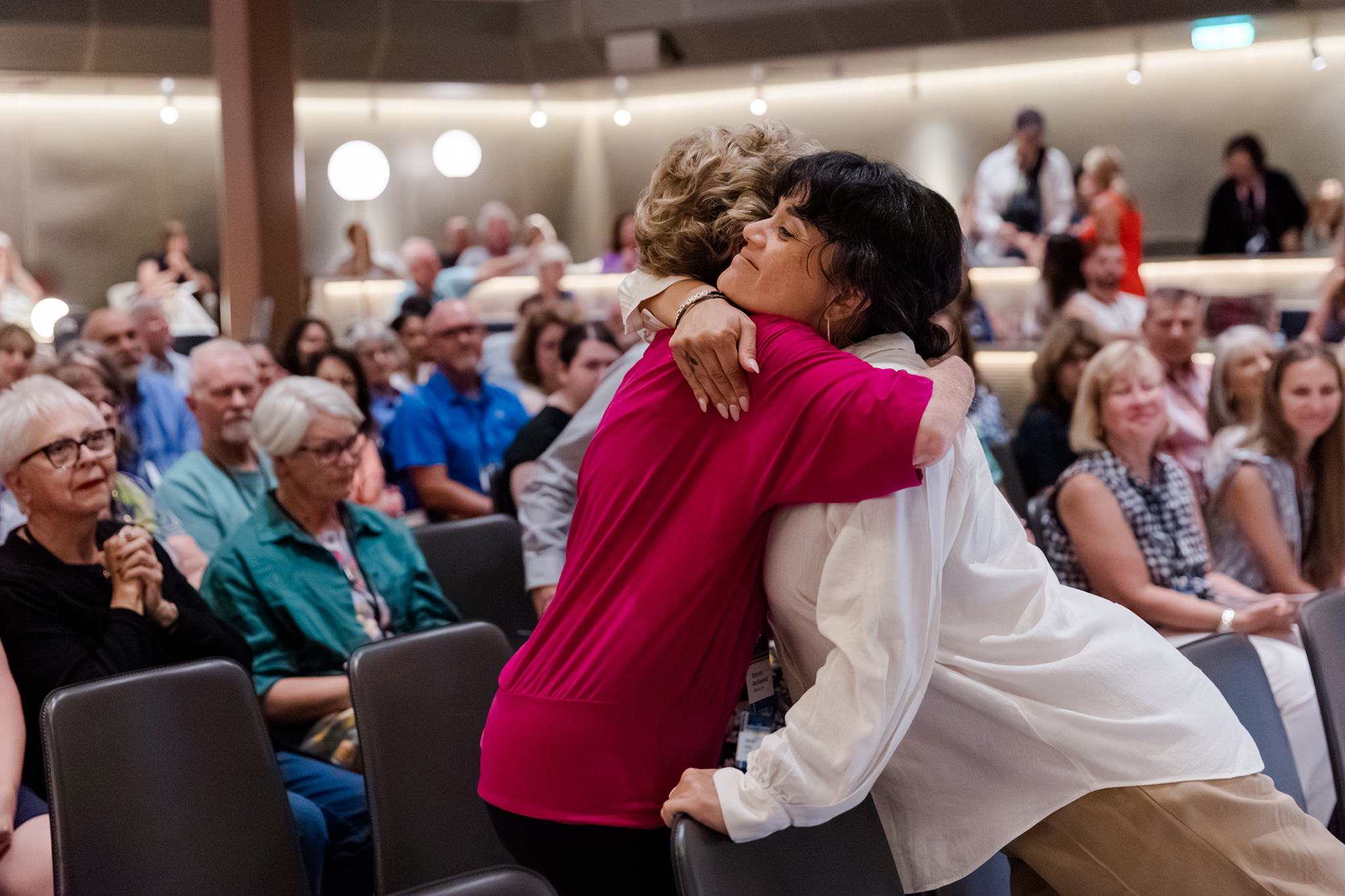 Two women share a warm embrace during a gathering, surrounded by seated attendees.