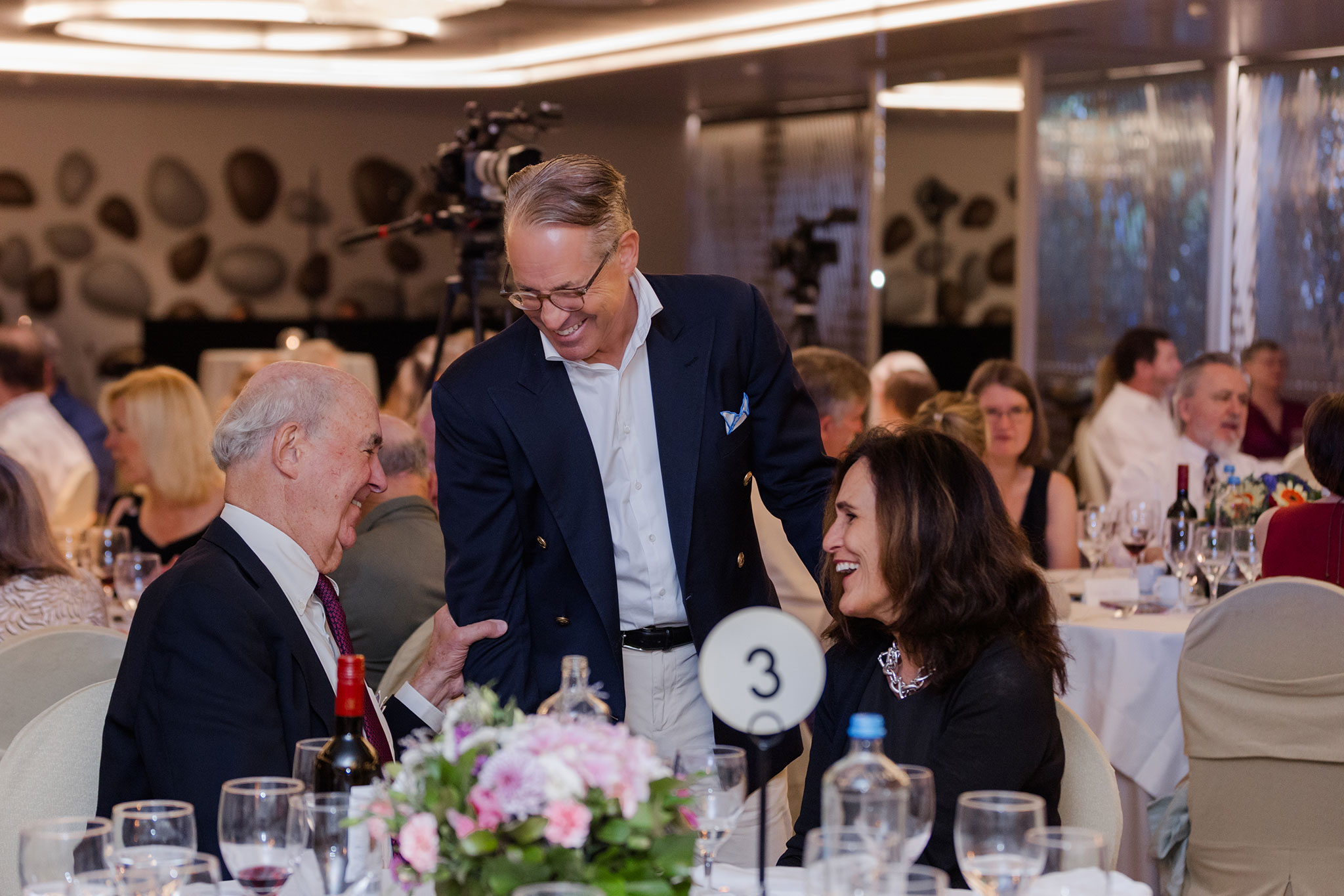 Eric Metaxas in navy blazer greets two smiling guests at a banquet dinner with wine glasses and a floral centerpiece.