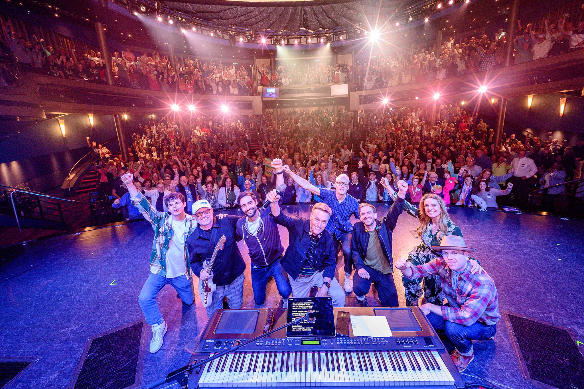 A band poses at the front of a stage with arms raised as a large audience cheers behind them in a packed theater.