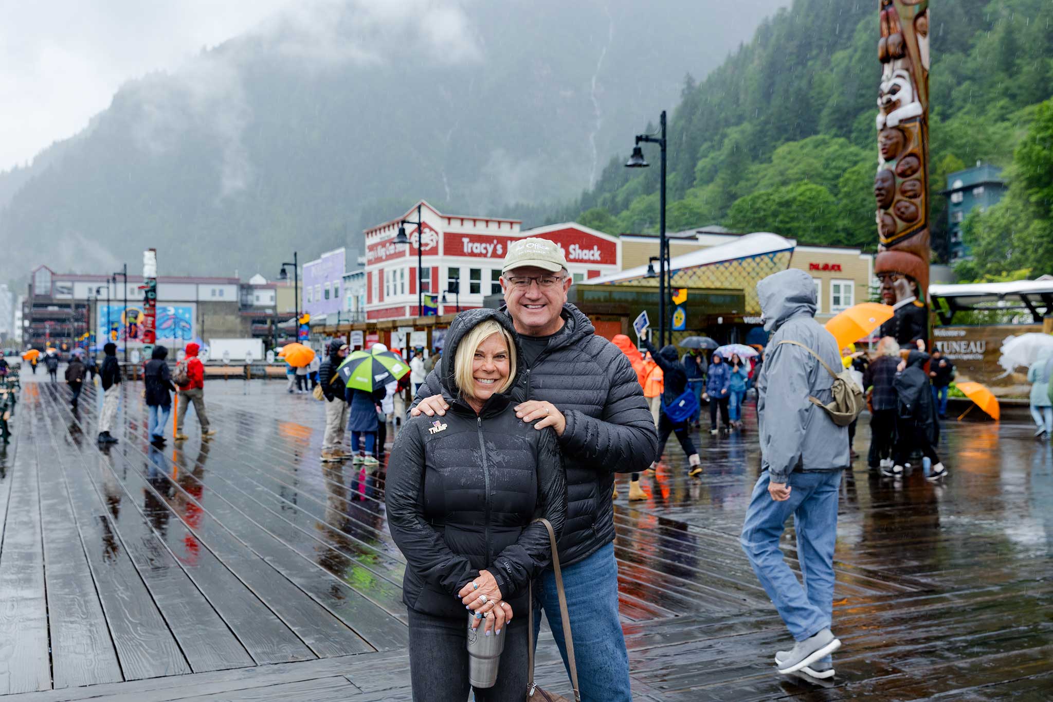 A couple stands together on a rainy day in Juneau, Alaska, as crowds with umbrellas walk the boardwalk lined with colorful storefronts.