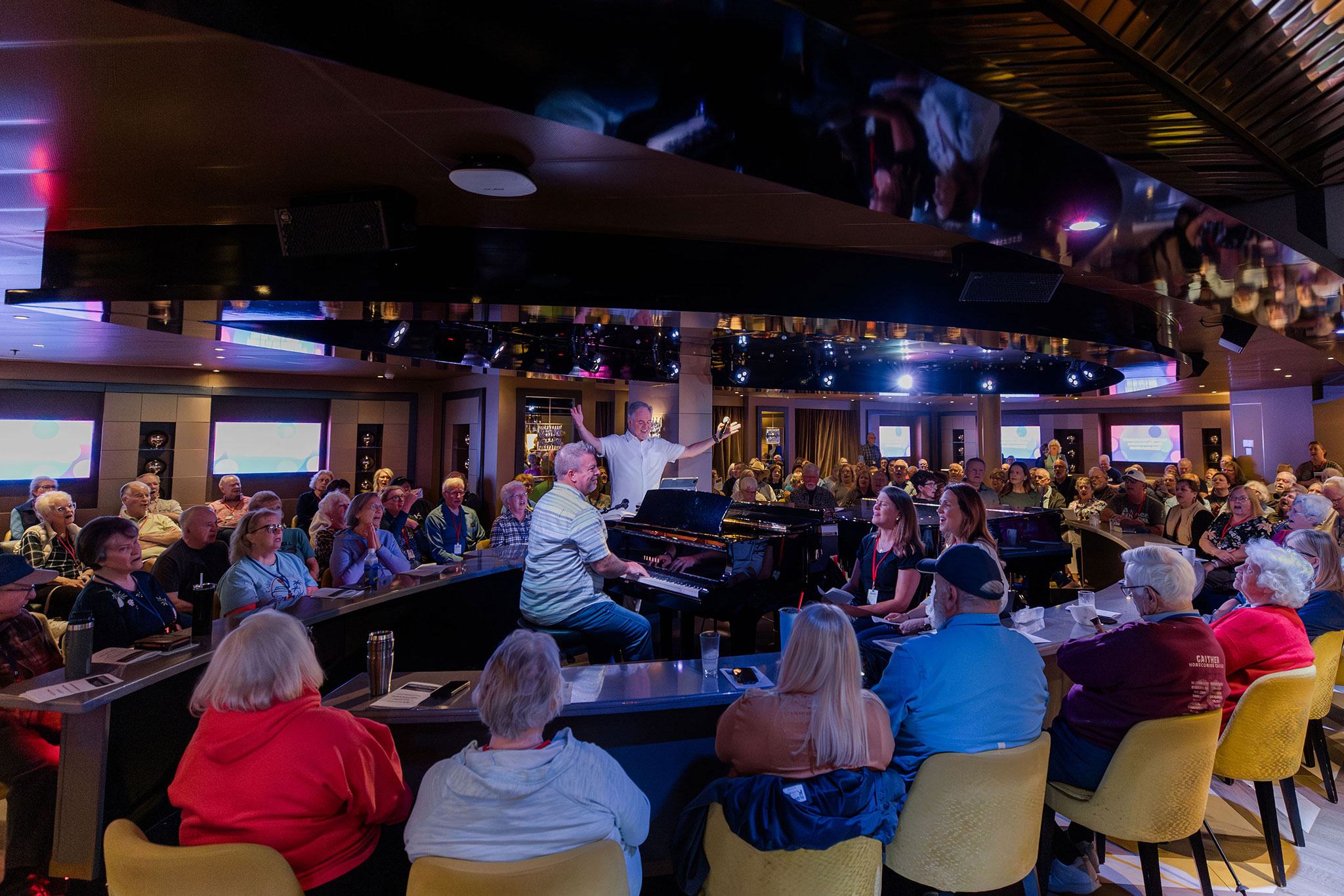 Cruise guests fill a lounge, seated around pianos as a musician leads an interactive sing-along session.