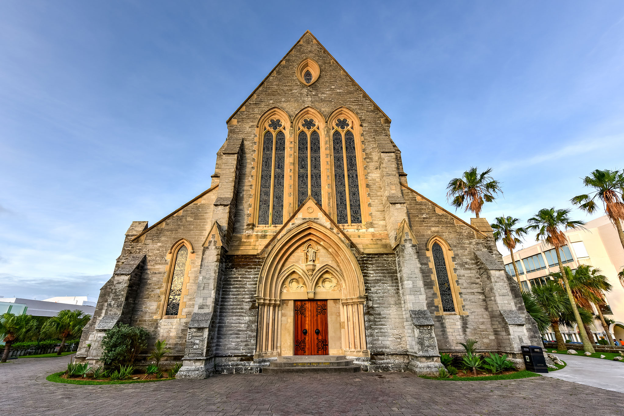 Gothic-style church facade glowing at sunset with stained-glass windows, towers and palms under a clear sky.