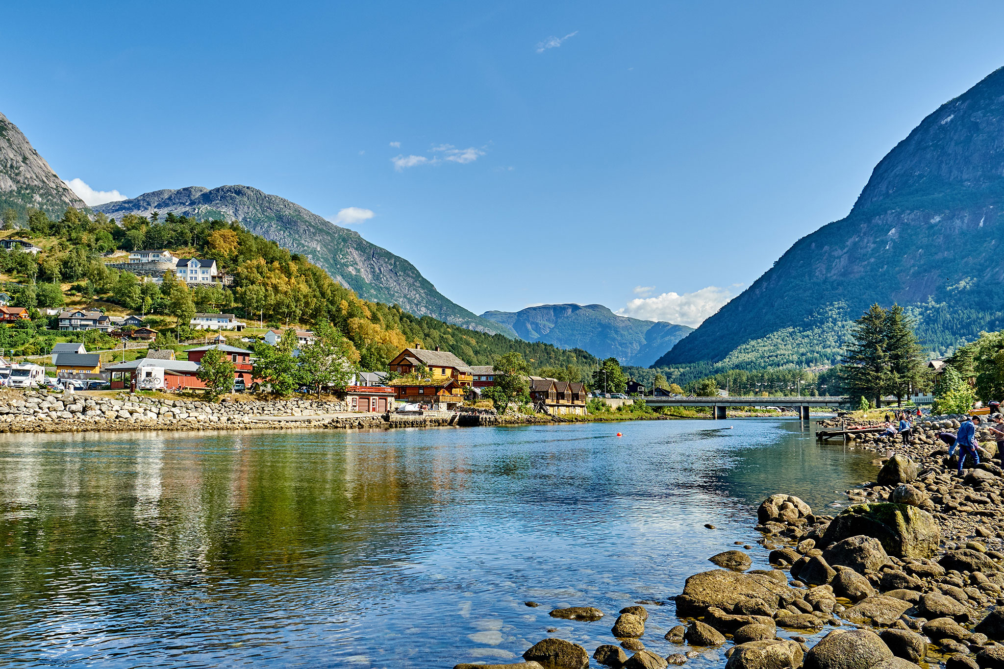 View of Eidfjord, Norway, with colorful houses along the riverbank, a stone bridge, and steep green mountains rising in the background.