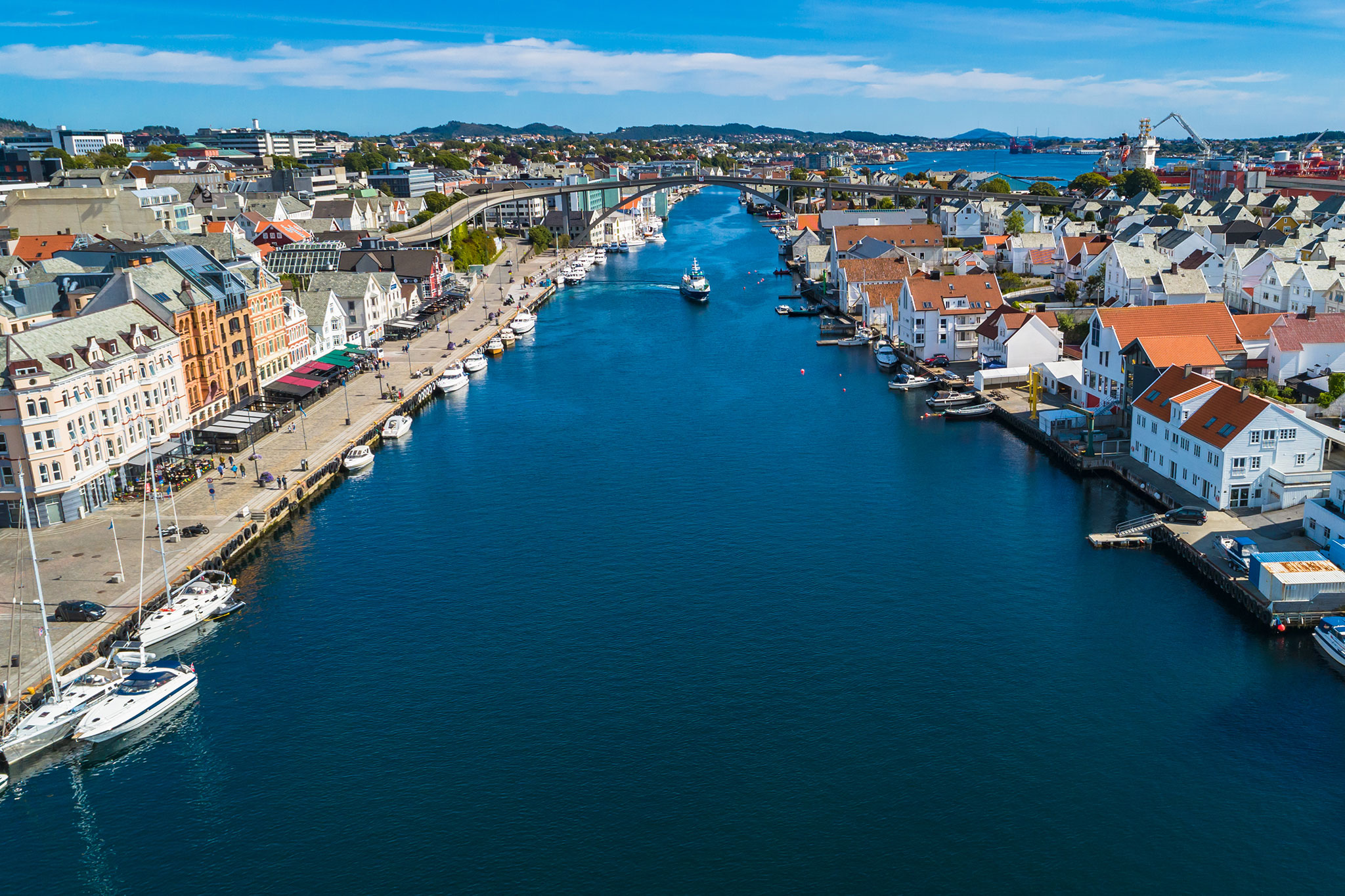 Scenic canal running through Haugesund lined with shops, cafes, marinas, and waterfront houses, with shipyards and cranes visible in the distance.