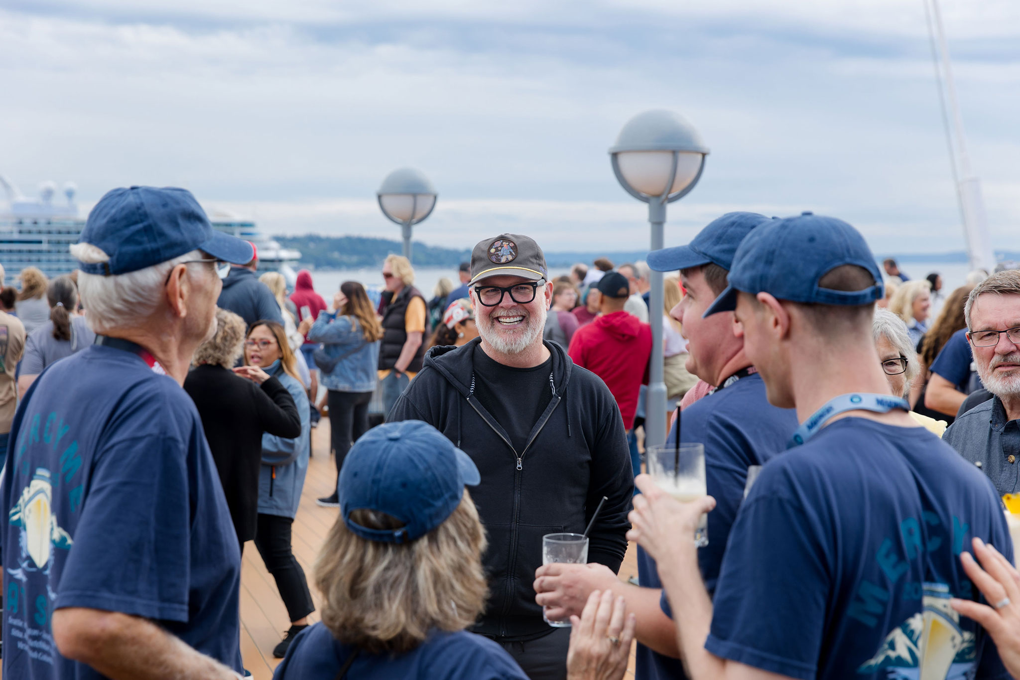 A smiling man in a black hoodie and cap chats with a group of people in blue shirts during a cruise deck gathering.