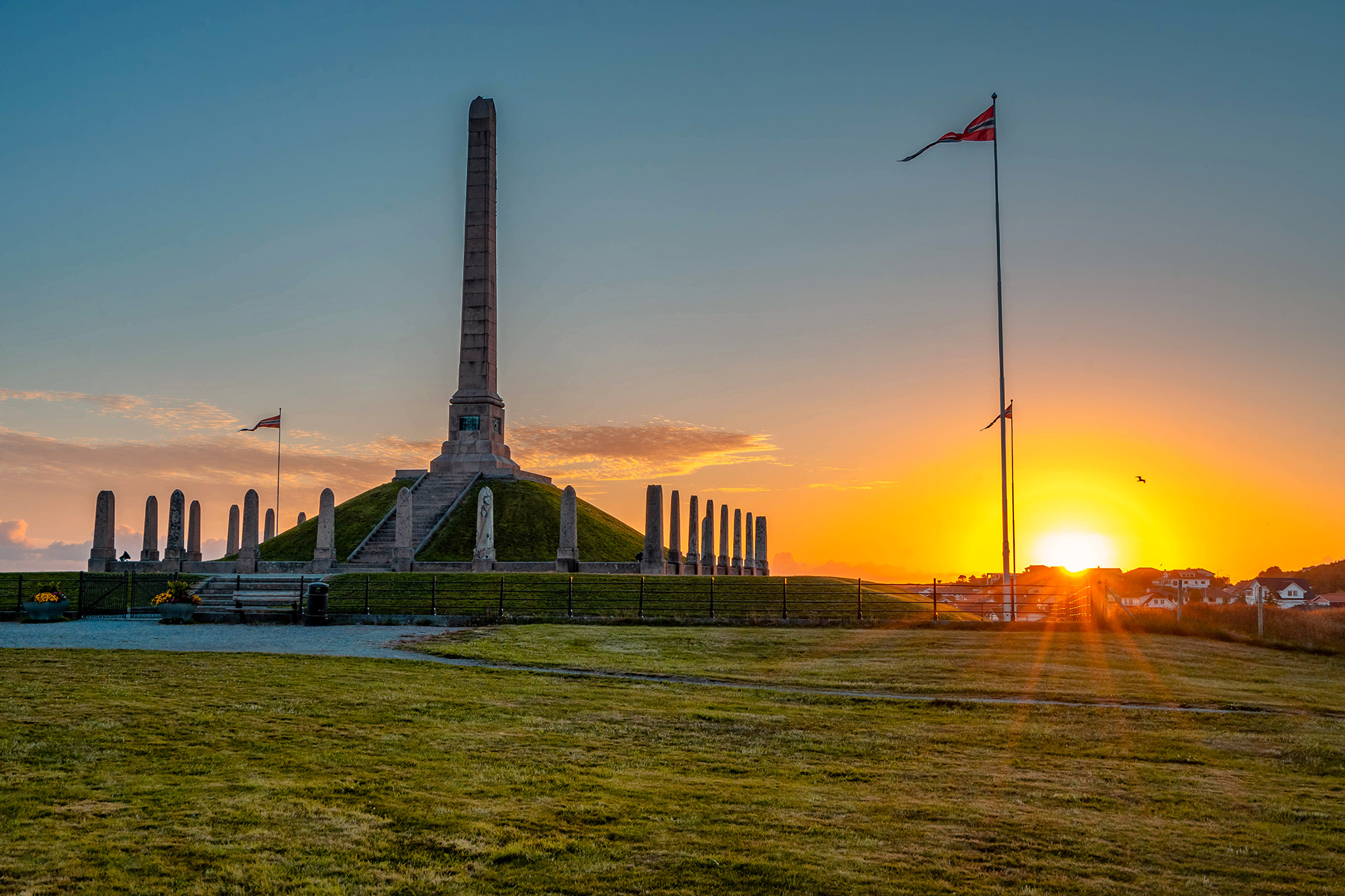Monument with tall stone obelisk and Norwegian flags at Haraldshaugen in Haugesund, Norway, illuminated by a golden sunset.