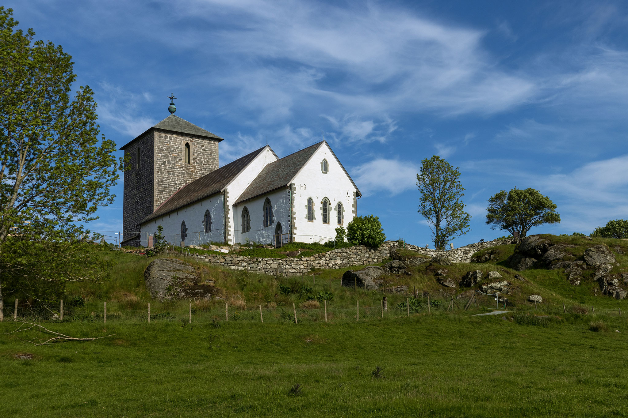 Historic white stone church with a tall tower sits on a grassy hillside surrounded by trees and rocks under a bright blue sky in Norway.