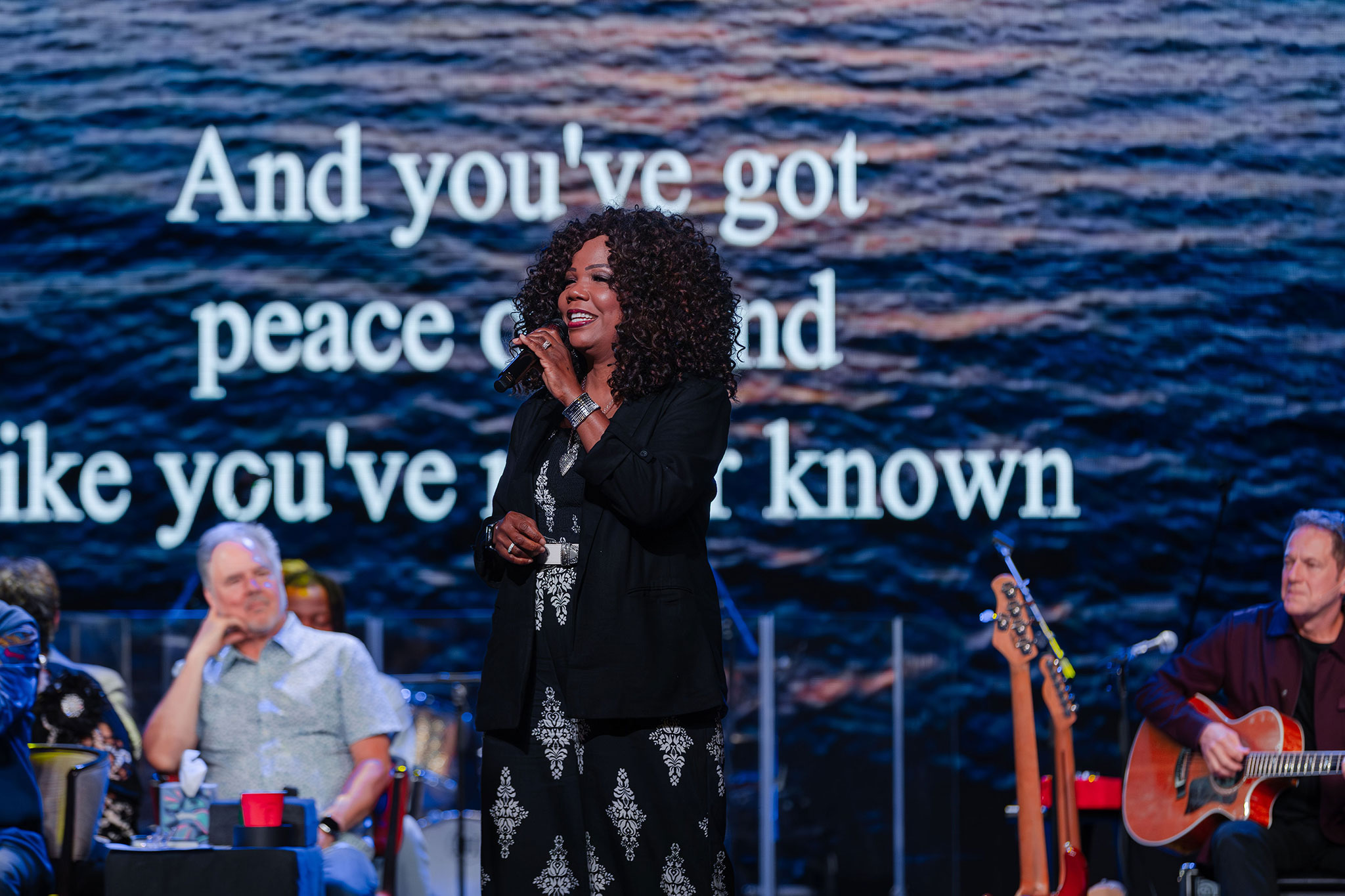 Lynda Randle sings onstage with a microphone in front of a large screen displaying song lyrics, while musicians and seated performers watch behind her.