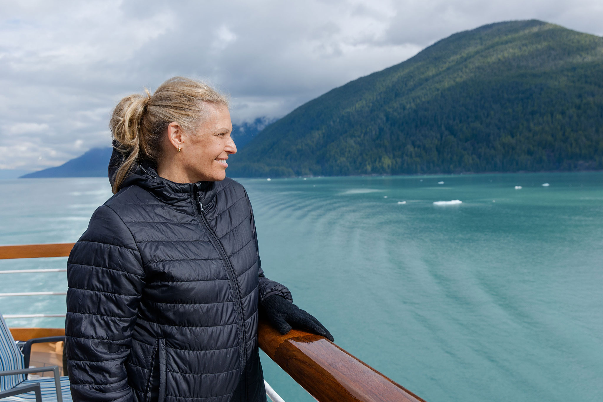 Woman in a black jacket stands on a ship’s deck, smiling as she looks out over calm turquoise water with forested mountain rising in the distance under a cloudy sky.