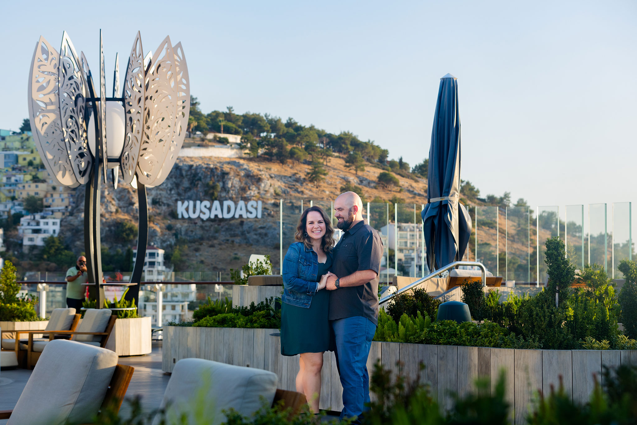 A couple stands together on an outdoor cruise ship deck, smiling and holding hands amid lounge seating and greenery. In the background, a hillside cityscape with the word “Kusadasi” is visible, indicating a port stop in Turkey.