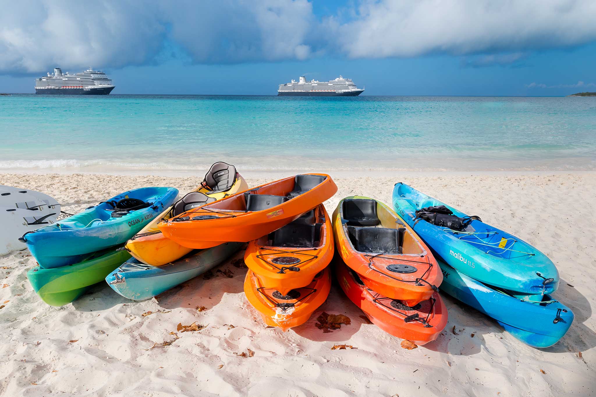 Colorful kayaks stacked on a sandy beach with turquoise ocean water in the foreground and two cruise ships anchored offshore beneath a partly cloudy blue sky.