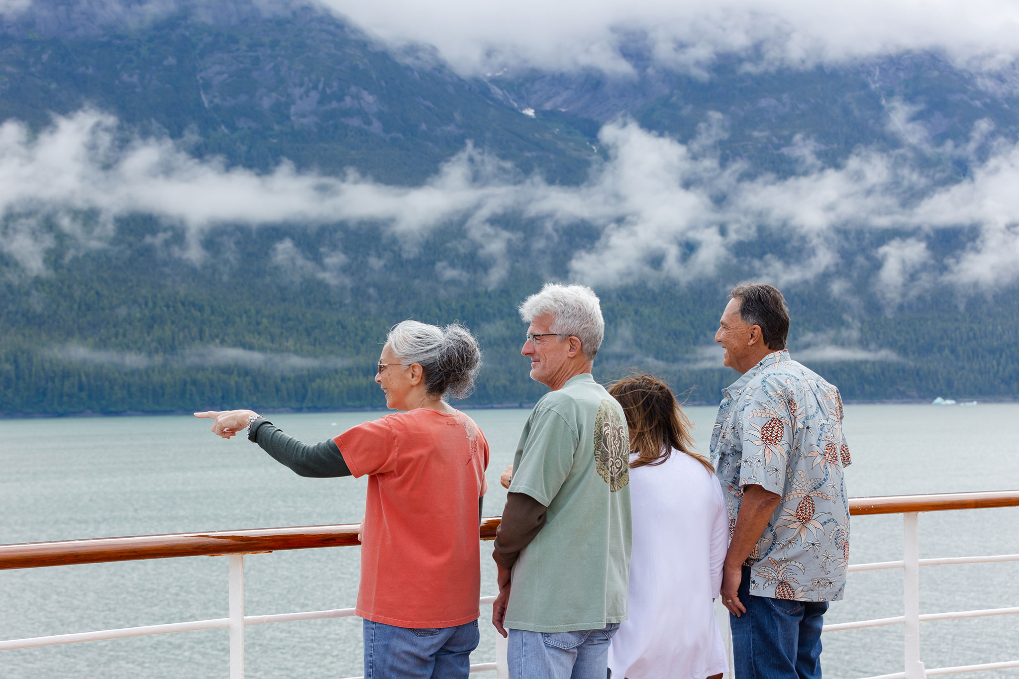 A group of travelers stands on a cruise ship deck in Alaska, looking out over calm water toward misty, forested mountains, with low clouds drifting across the landscape.
