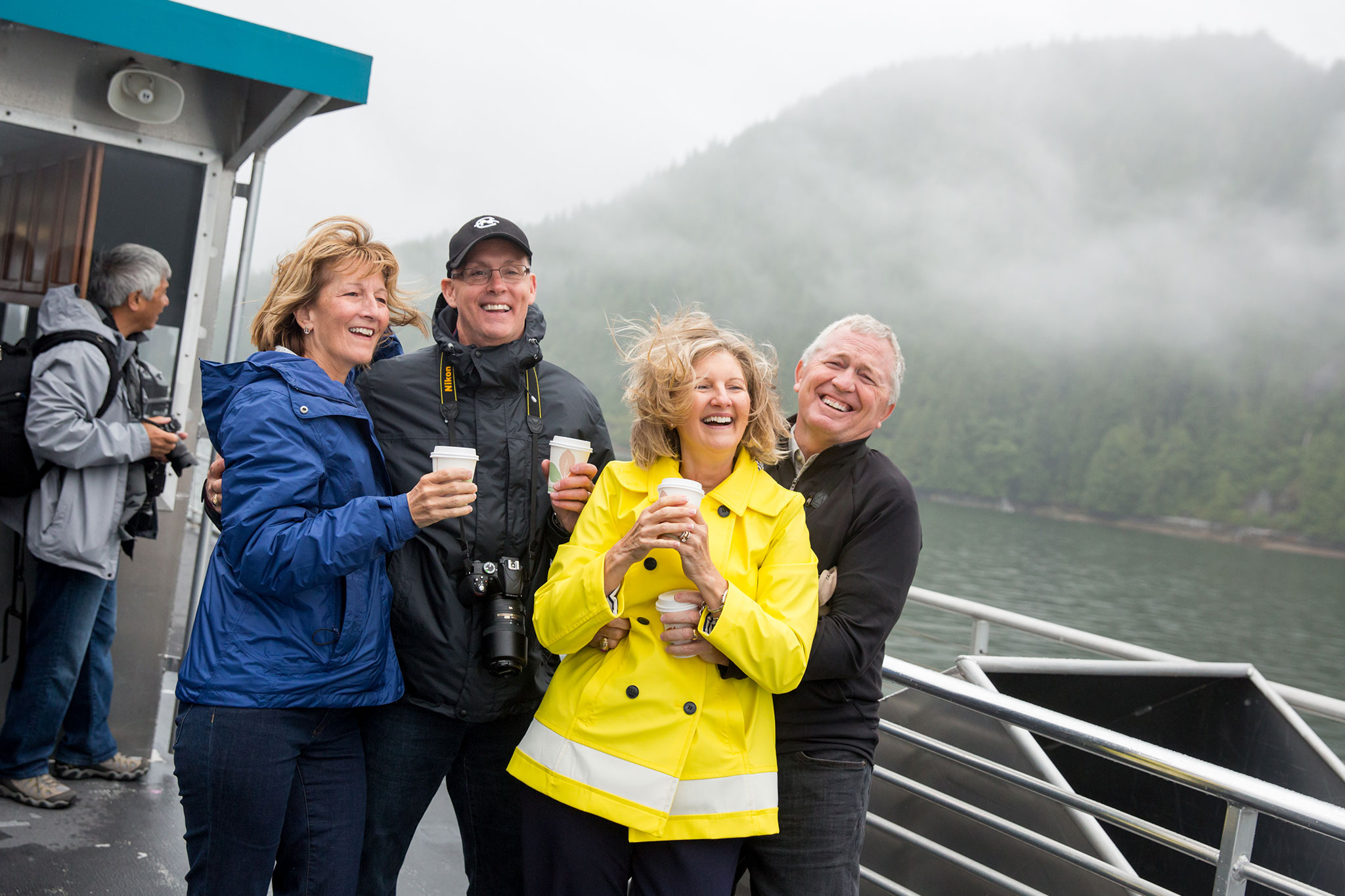 A group of travelers bundled in jackets smile while holding warm drinks on an outdoor boat deck in Alaska, with misty forested mountains and calm water in the background.