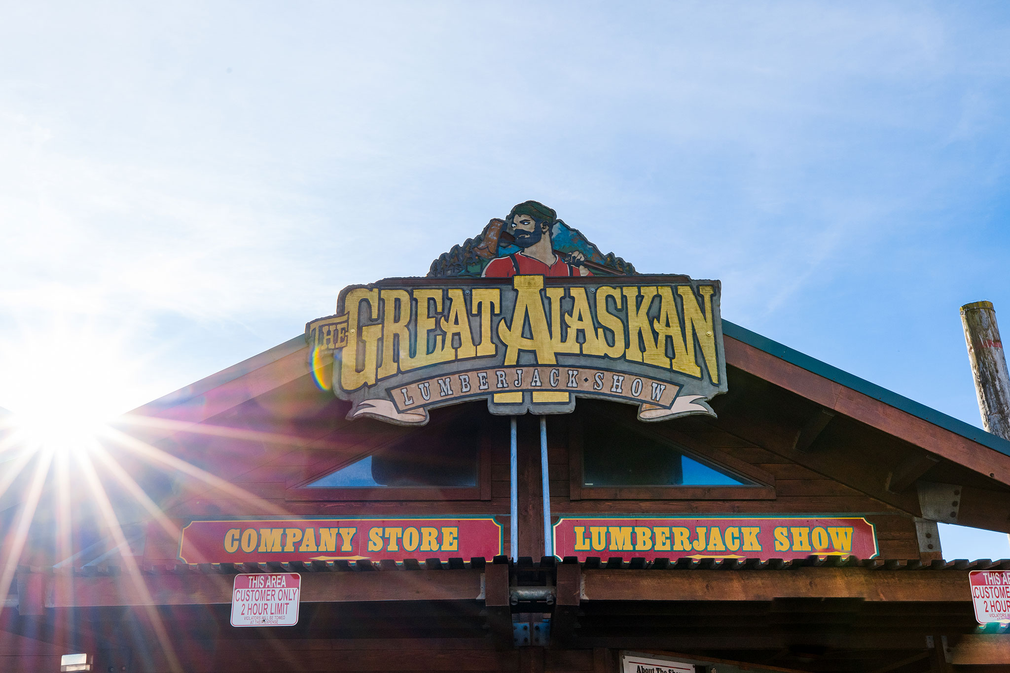 A sign for the Great Alaskan Lumberjack Show in Ketchikan, Alaska, is mounted on a rustic wooden building with bold lettering, a lumberjack illustration above, and sun rays shining across a clear blue sky.