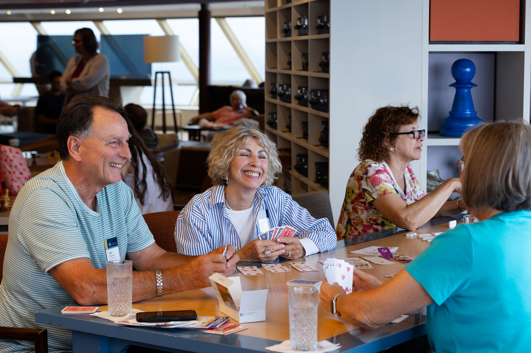 A group of travelers laugh and play cards together in a bright lounge onboard a cruise ship, with large windows, shelves, and other guests relaxing in the background.