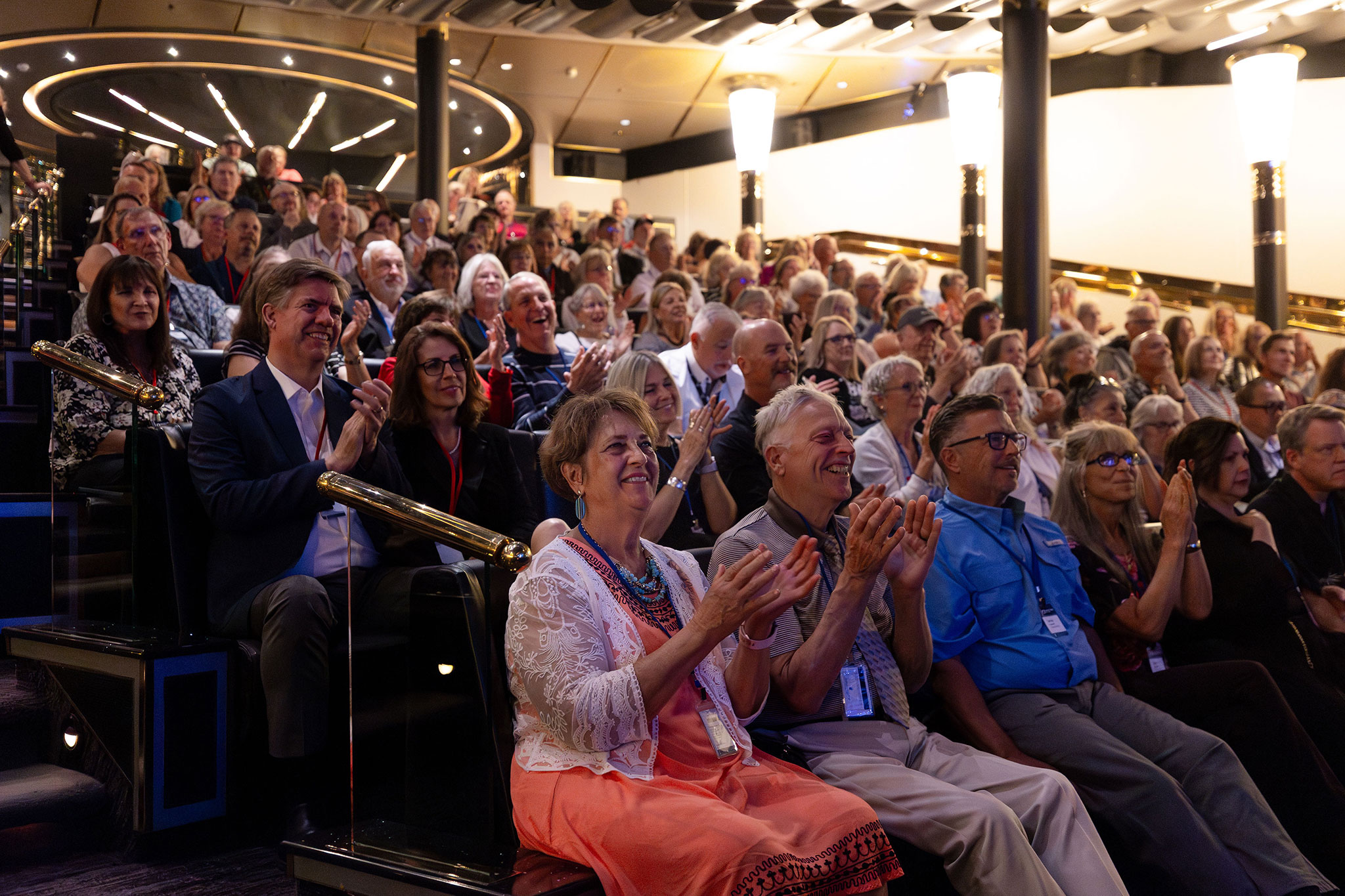 An audience of travelers seated in a cruise ship theater smile and applaud during a live presentation, with warm lighting and tiered seating throughout the venue.
