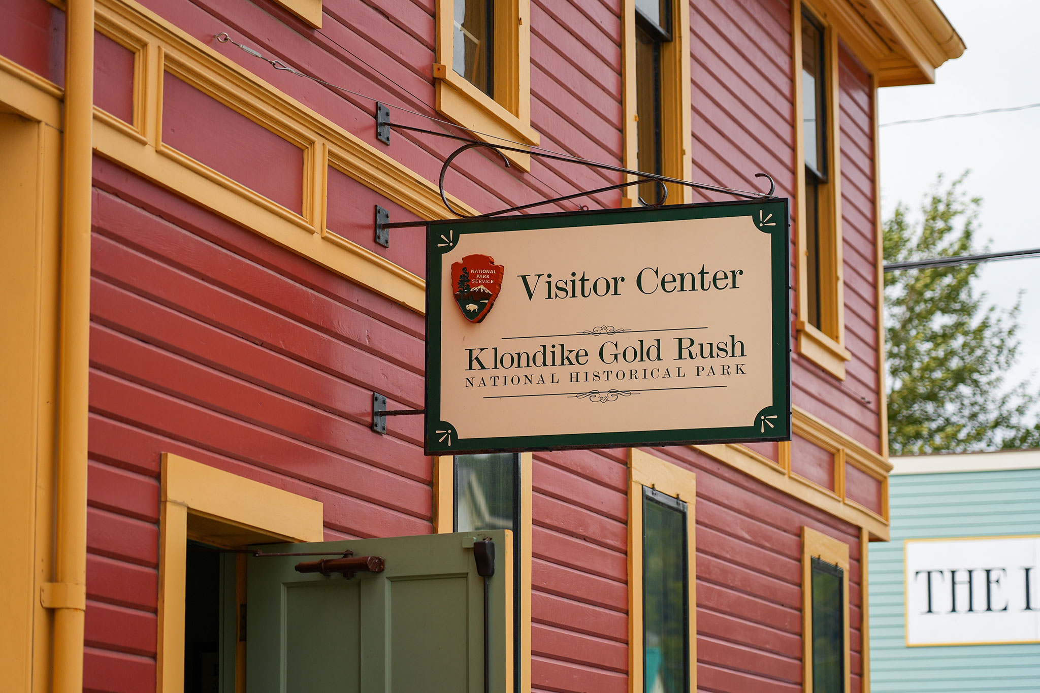 Close-up of a hanging sign reading “Visitor Center Klondike Gold Rush National Historical Park” mounted on a red wooden building with yellow trim in Skagway, Alaska; there is a partially open green door below the sign.