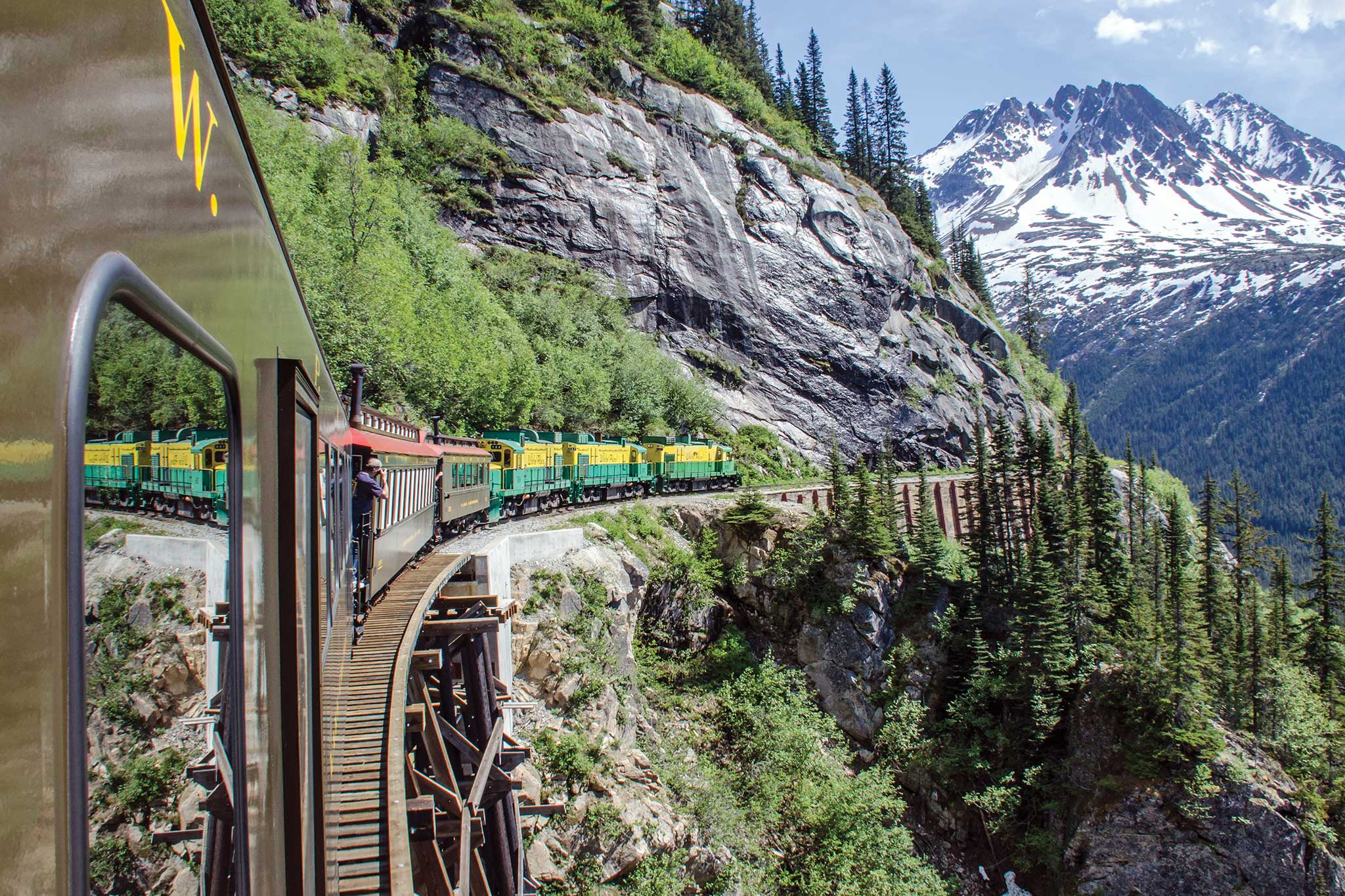 View of the White Pass & Yukon Route Scenic Railway as the train curves along a narrow mountainside track in Alaska, crossing a wooden trestle above a rocky gorge, with evergreen trees, cliffs and snow-capped peaks rising in the distance.