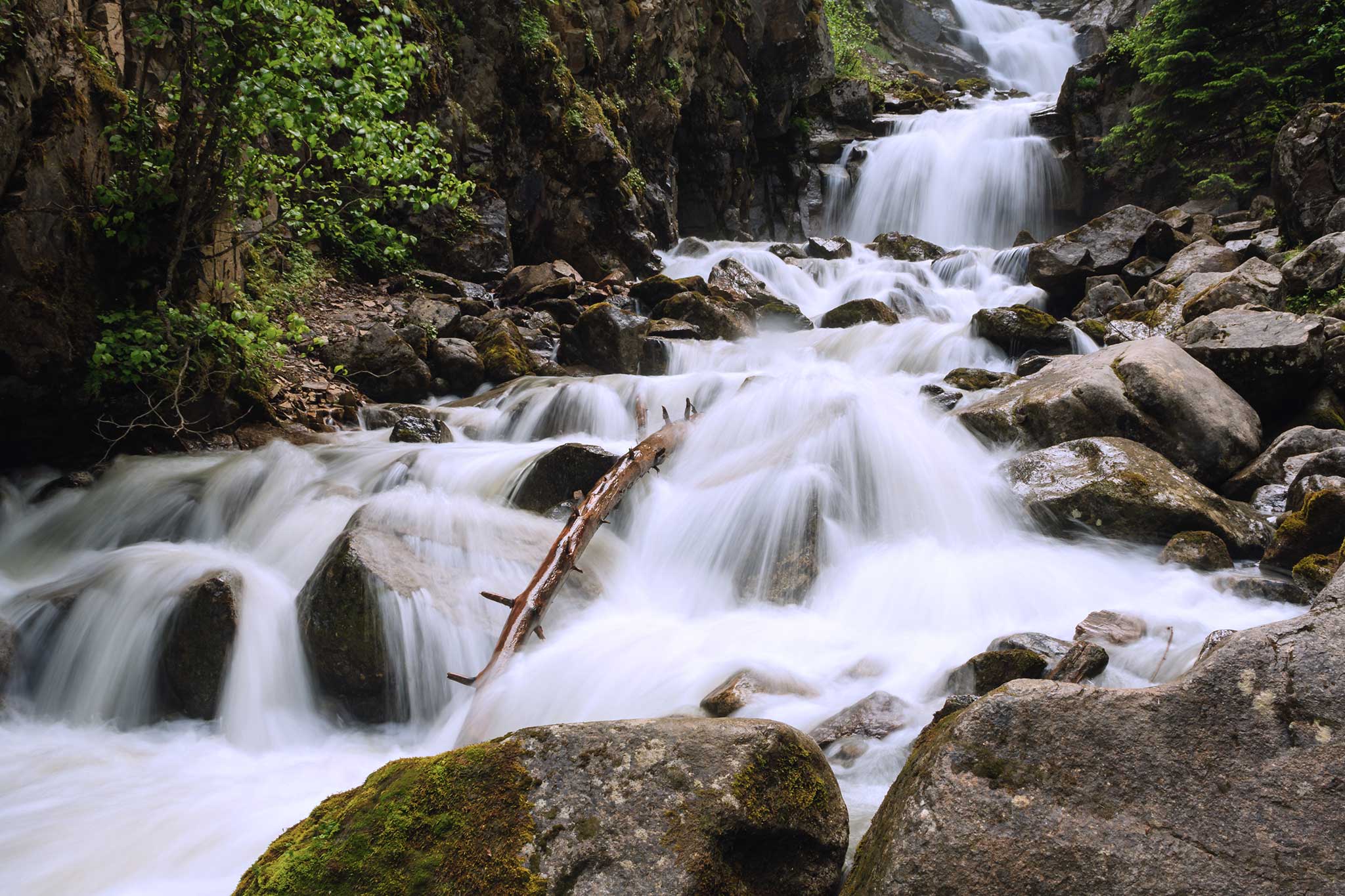 A cascading waterfall flows over smooth rocks in a lush forest setting, with white rushing water weaving around moss-covered stones and greenery along the rocky banks.