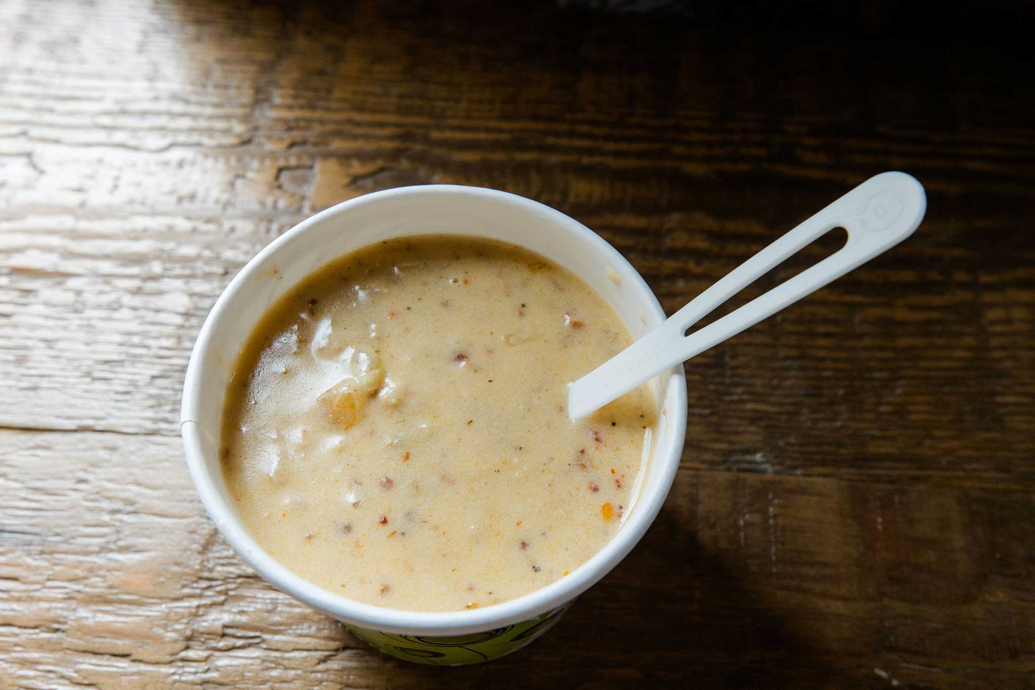 A cup of creamy clam chowder sits on a rustic wooden table, with a white plastic spoon resting inside the bowl of thick, hearty soup.