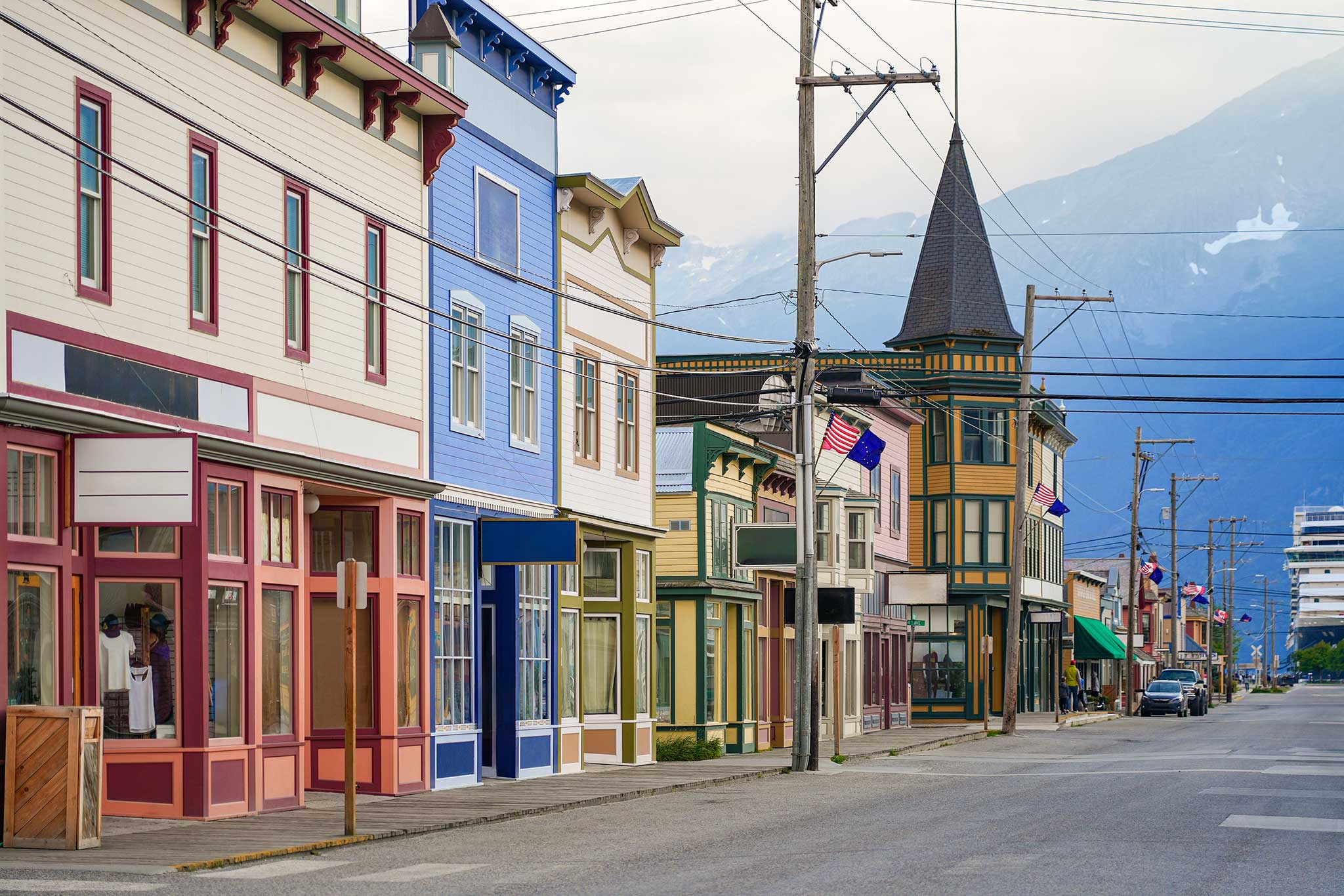 Colorful historic storefronts line a quiet street in downtown Skagway, Alaska, featuring pastel-painted buildings, a turreted corner structure with flags, and mountains rising in the distance.