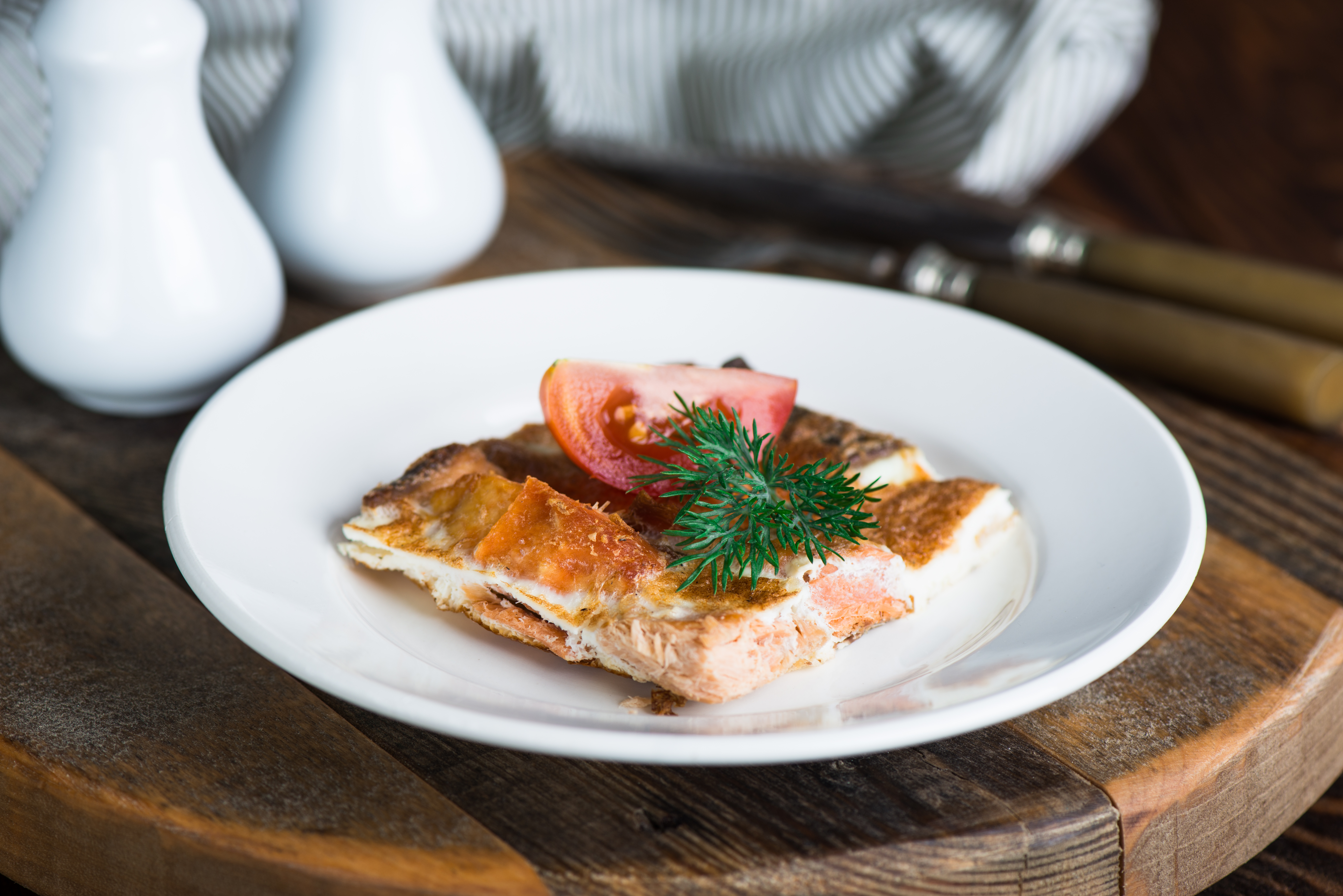 Grilled salmon fillet on a white plate garnished with fresh parsley and tomato, served on a rustic wooden table.