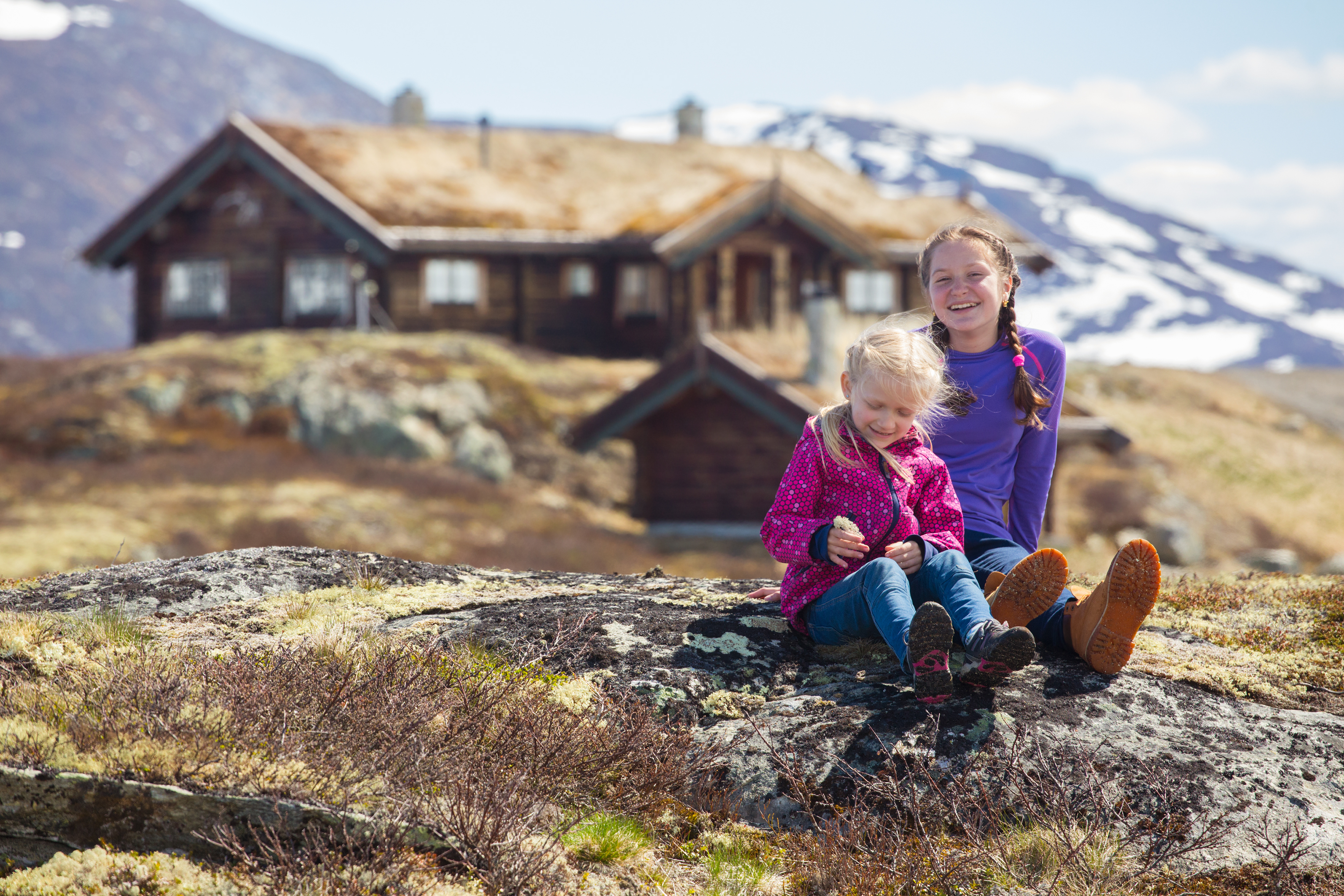 Two children sitting on rocky terrain near a rustic mountain cabin in Norway, with snow-covered peaks in the background.
