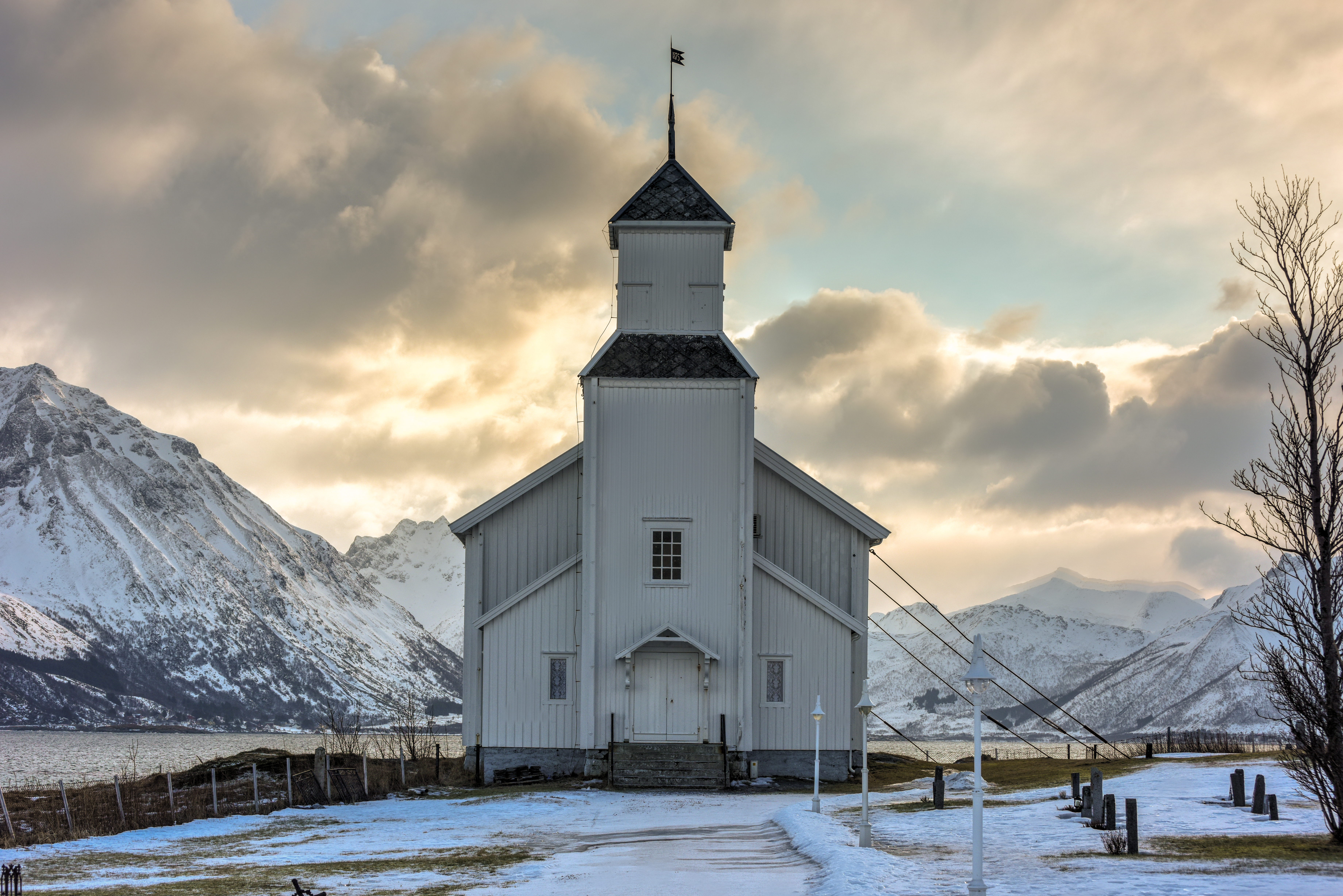 Gimsoy Church, a historic white wooden parish church in the Lofoten archipelago, Norway, set against snow-covered mountains and a coastal fjord landscape.