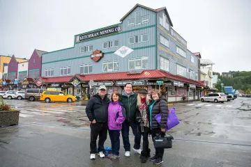 Four people smiling in front of Ketchikan Mining Co. and colorful shops on a rainy day