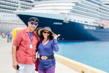 A smiling couple in vacation attire poses on a sunny cruise dock with large ships in the background.