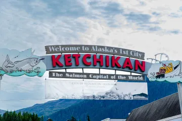 The welcome sign for Ketchikan, Alaska, reading “Welcome to Alaska’s First City—Ketchikan, the Salmon Capital of the World,” with colorful fishing-themed illustrations, is set against a backdrop of forested mountains and a cloudy sky.