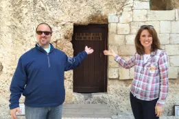 Man and woman smiling in front of the Garden Tomb in Jerusalem, pointing to the sign that reads “He is not here—for He is risen.”