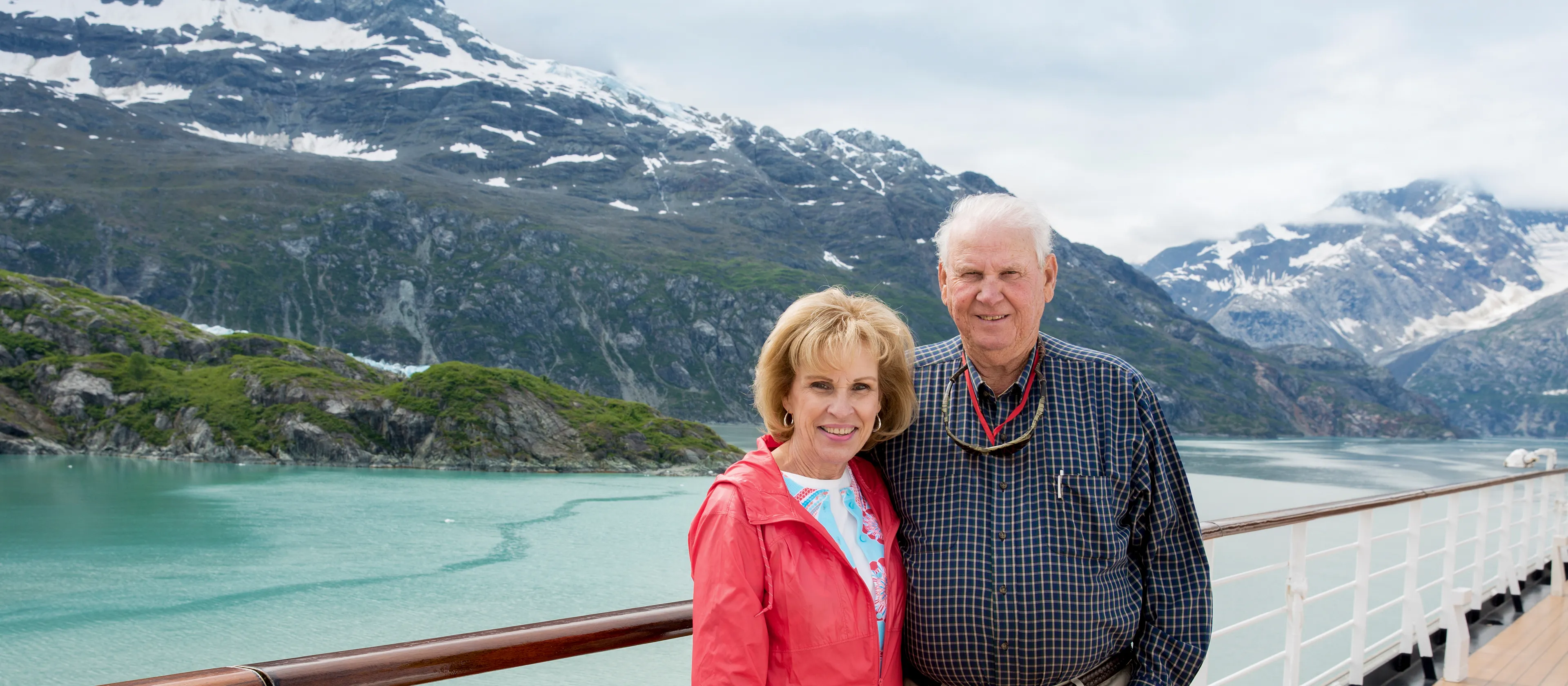 Smiling couple standing on the cruise ship deck with blue water and snowcapped mountains in the background