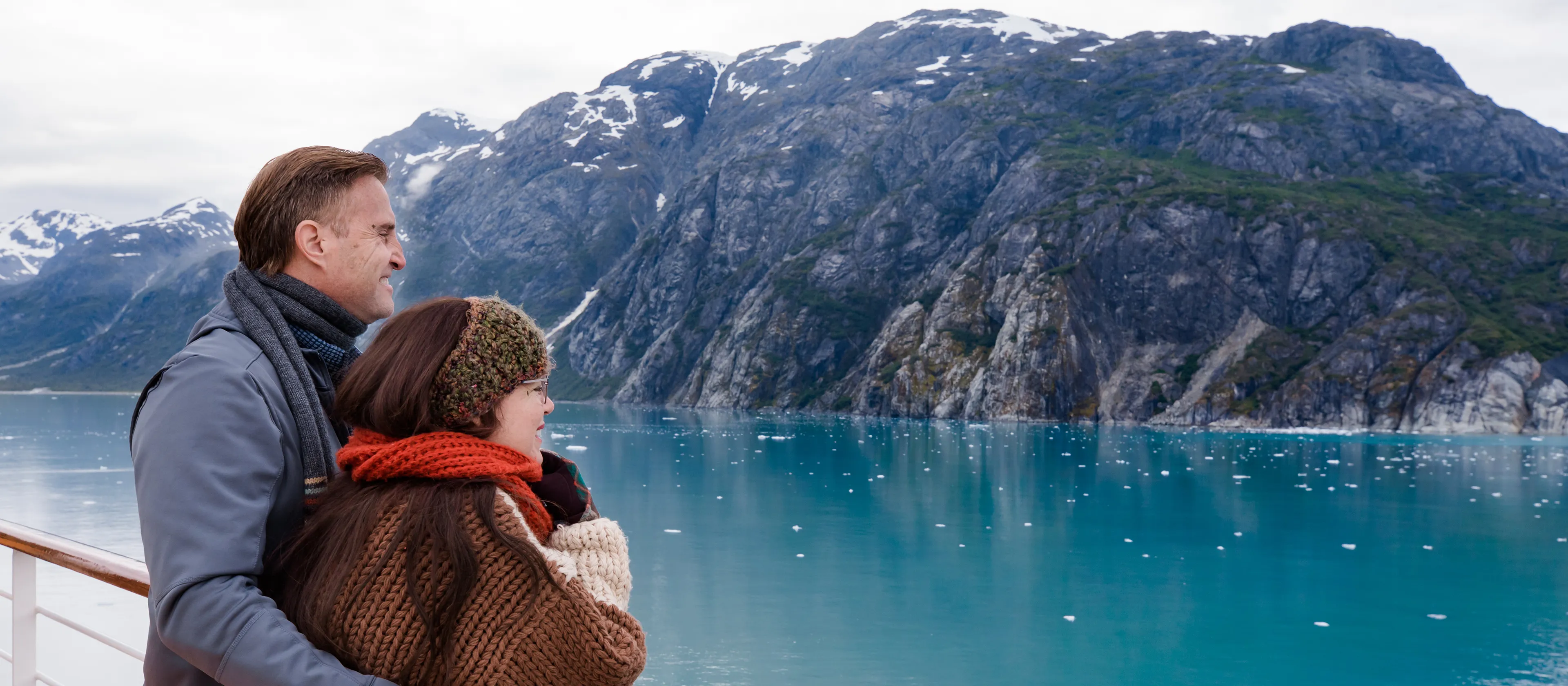 Smiling couple standing on the cruise ship deck with blue water and snowcapped mountains in the background